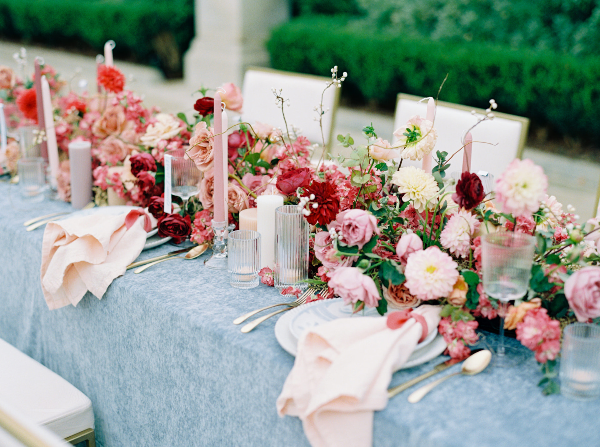 Garden-inspired wedding reception tablescape with pink and burgundy floral arrangements, candlelight, and soft blue linens by EVENT by OVA.