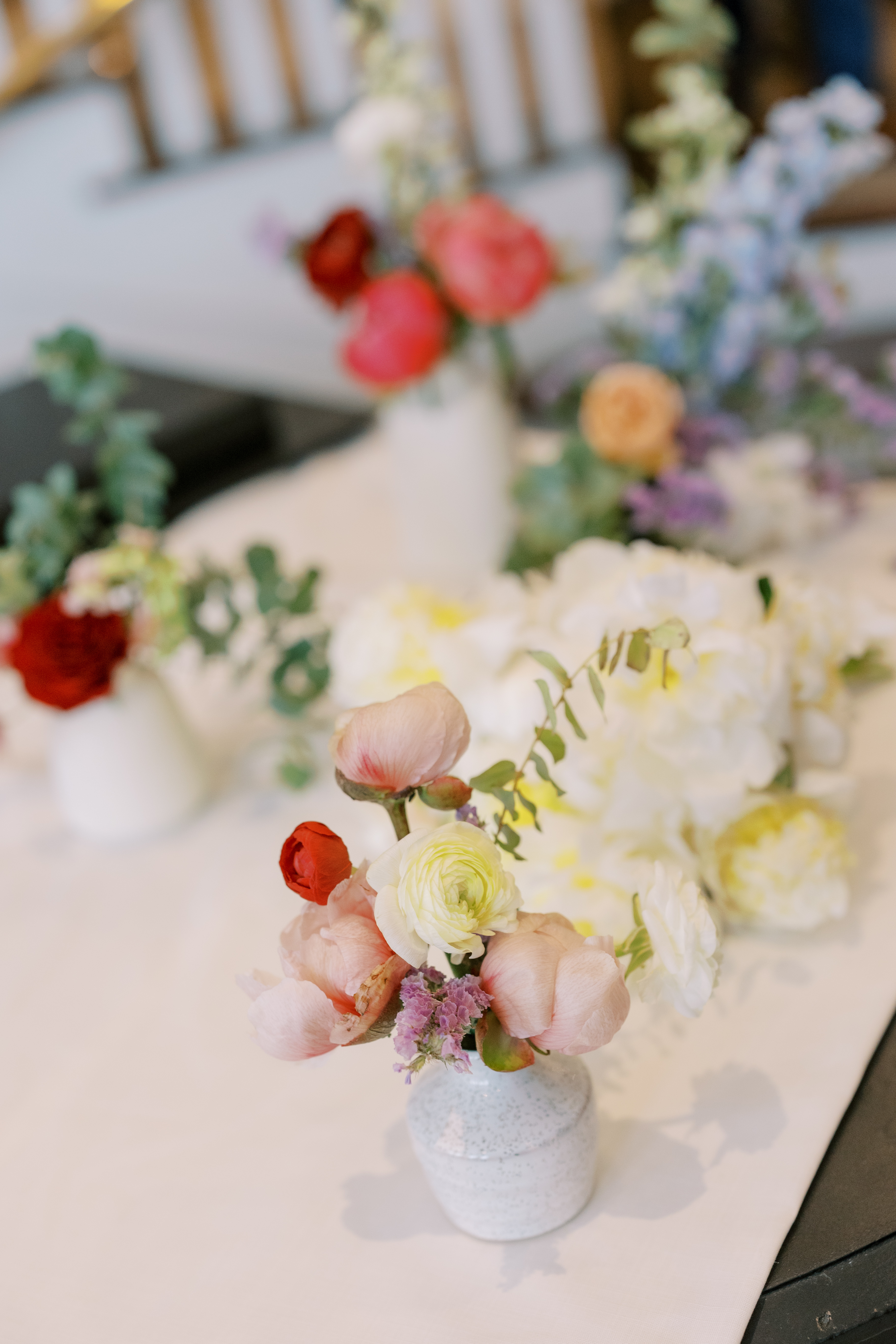 A speckled ceramic bud vase filled with pale pink peonies, creamy white ranunculus, and delicate purple accents on a reception table.