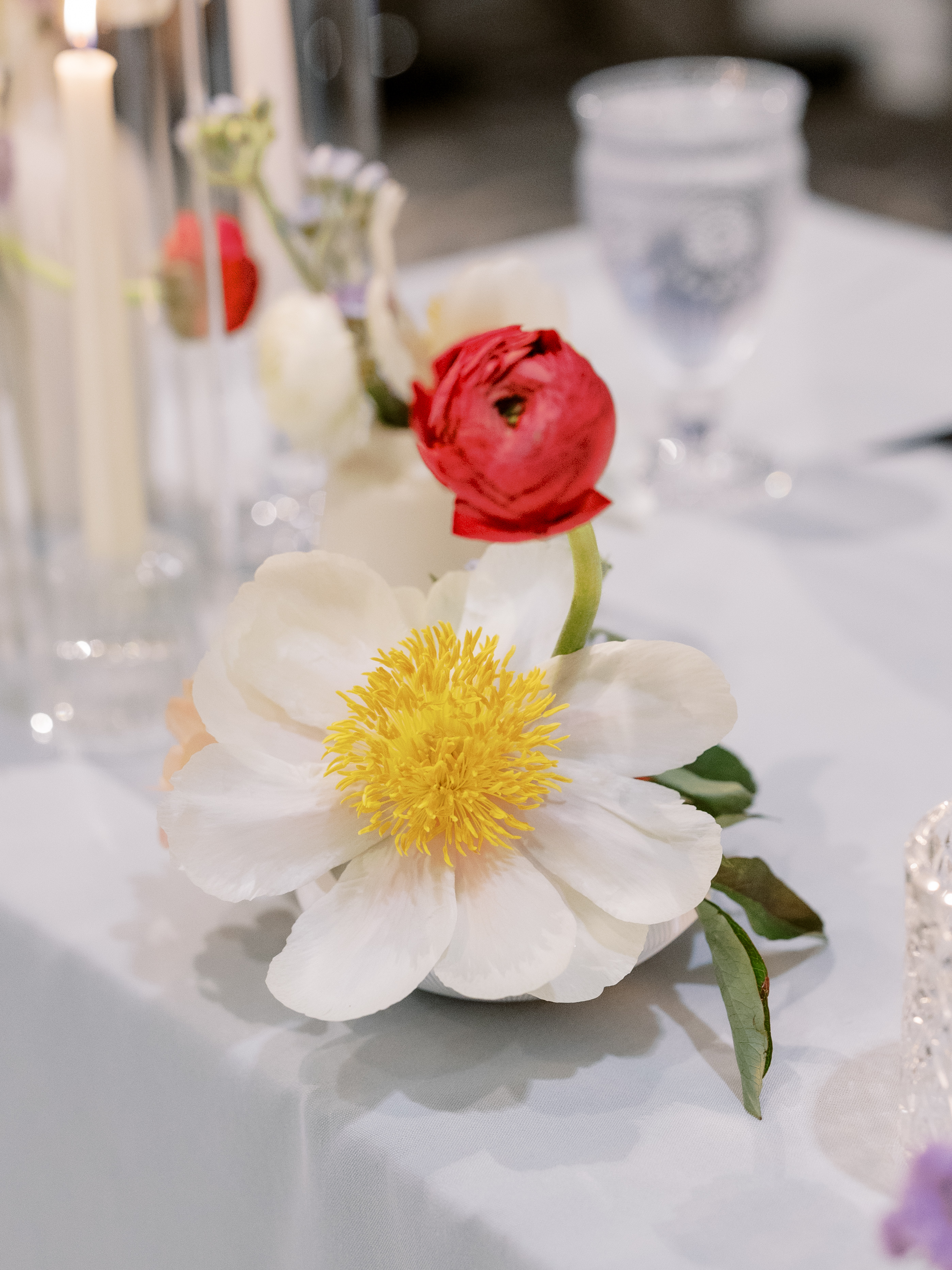 A close-up of an open white peony with a bright yellow center paired with a vibrant red ranunculus bloom on a reception table.