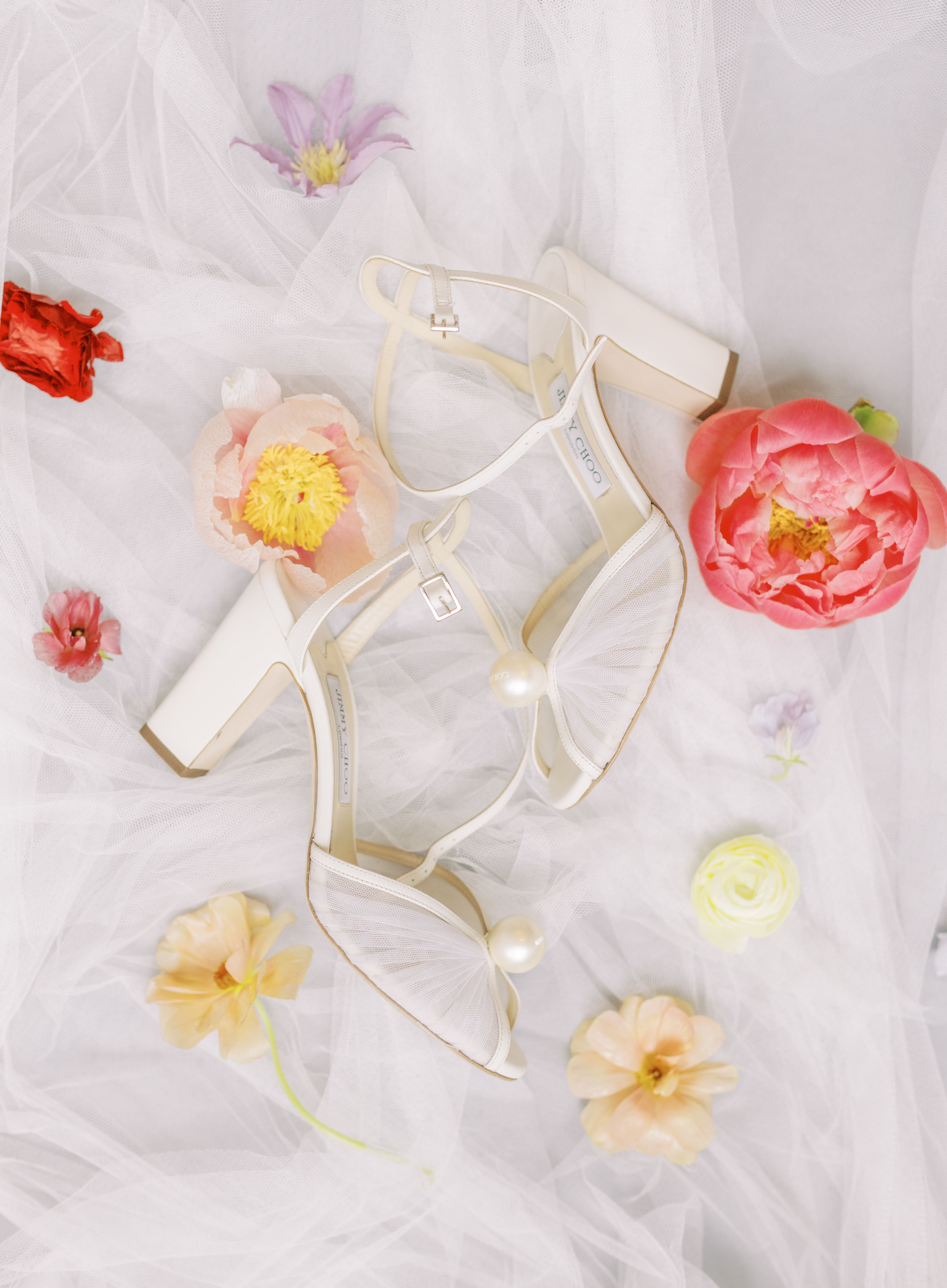 Elegant white bridal sandals adorned with large pearl embellishments resting on draped white tulle and soft floral petals.