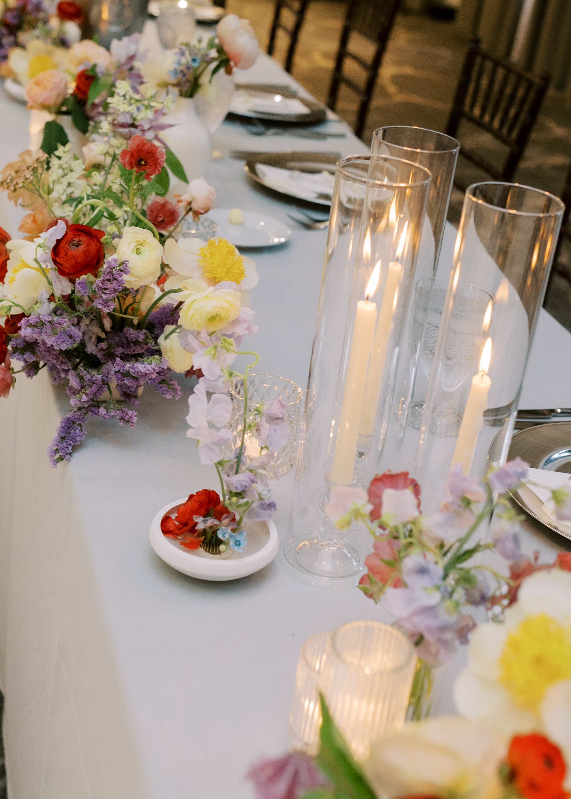 Lit taper candles in glass hurricane vases and delicate pastel floral arrangements on an elegant reception table.