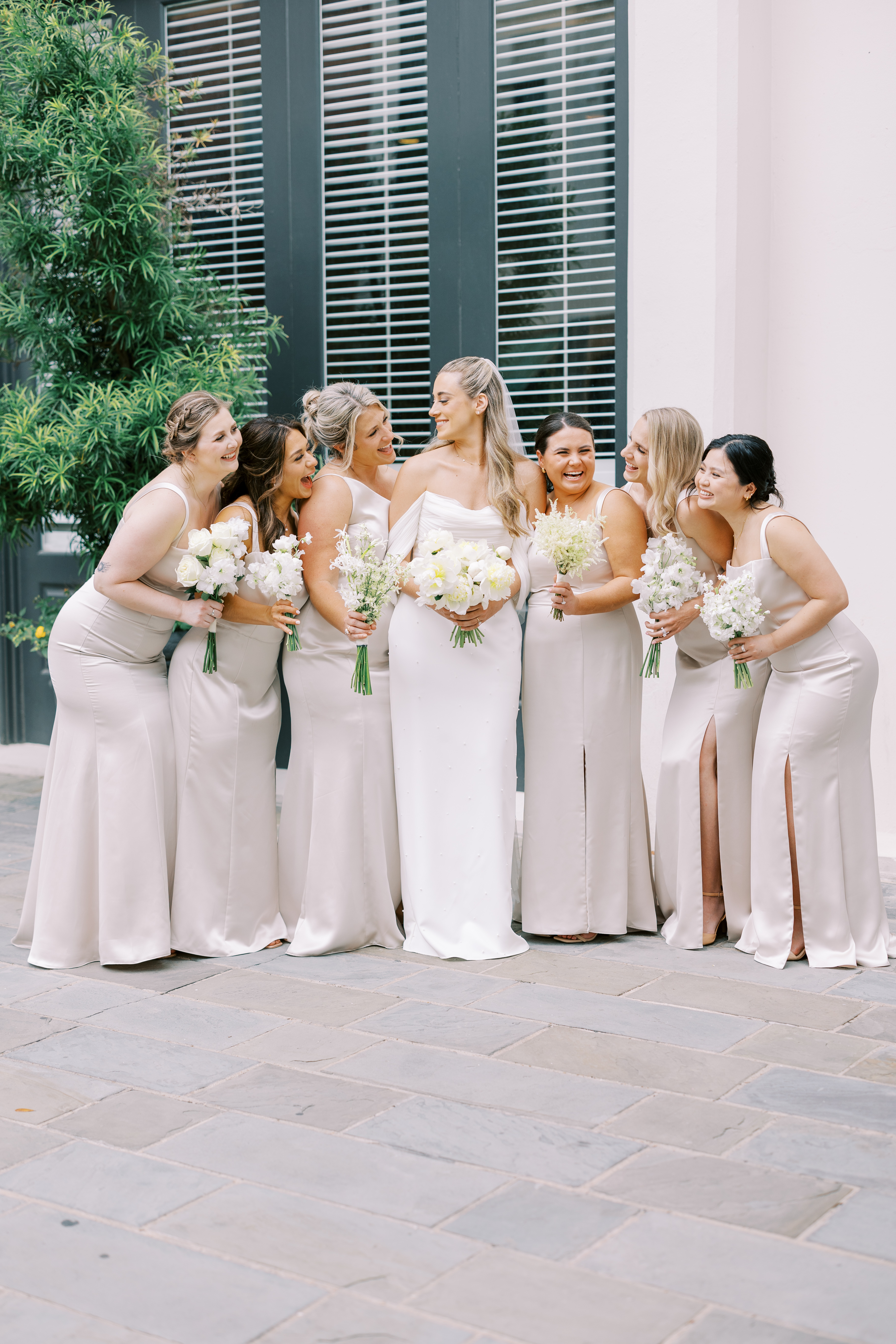 A bride smiles with bridesmaids in champagne dresses holding delicate white floral bouquets against louvered windows.