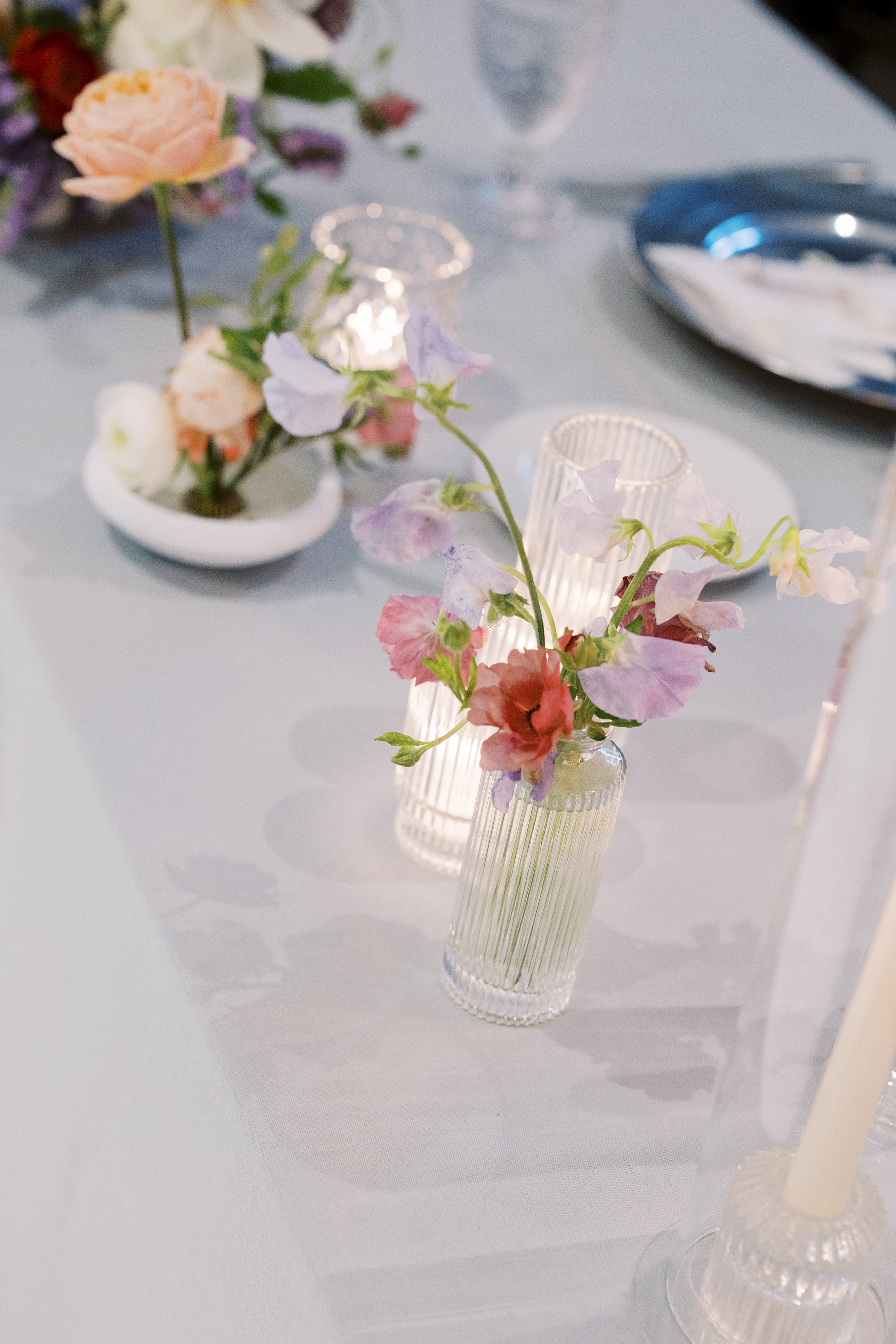 Delicate lavender sweet peas and pink blossoms arranged in fluted glass bud vases beside glowing candlelight on a reception table.