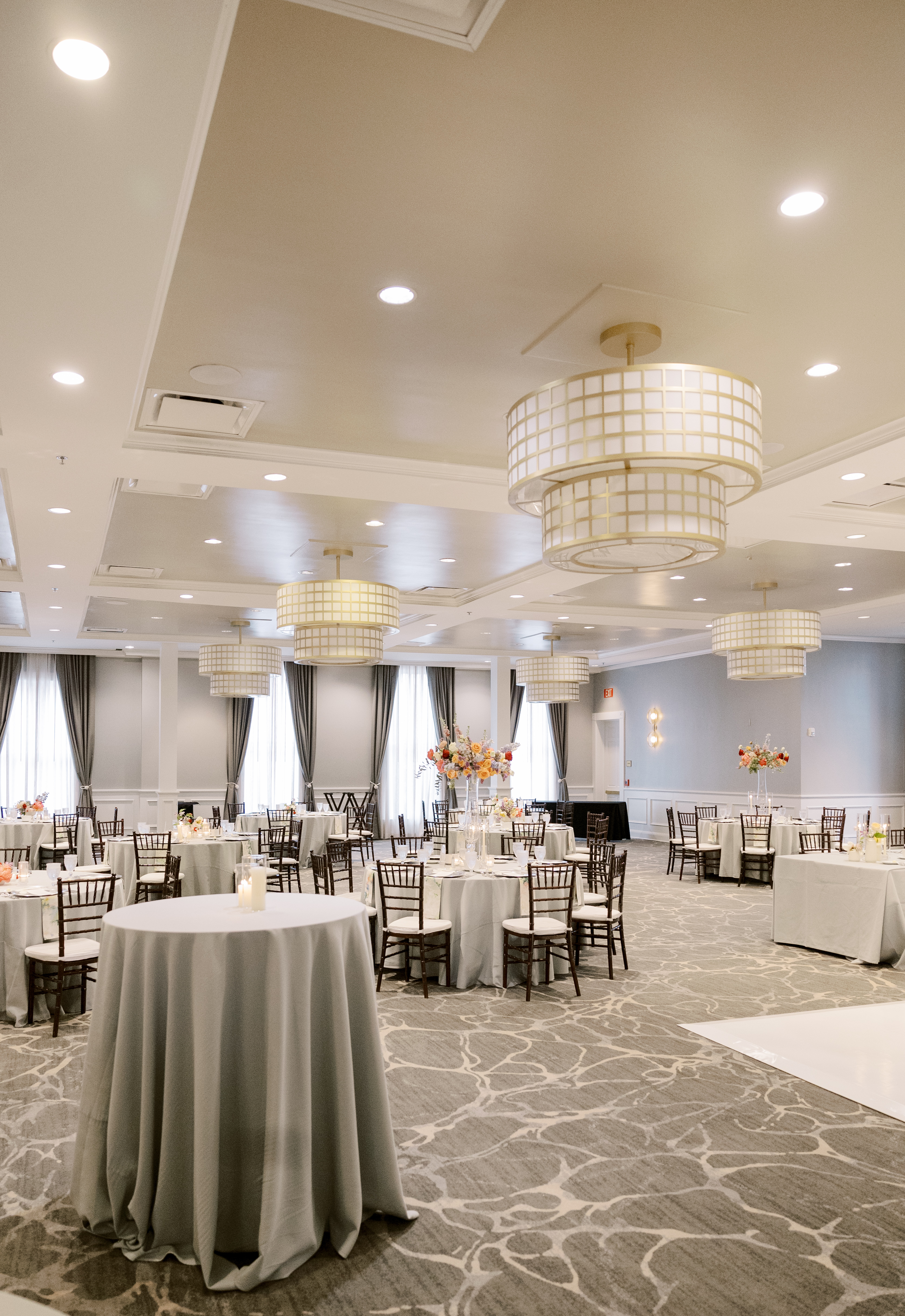 A ballroom reception setting with round dining tables, Chiavari chairs, and tall pastel floral centerpieces under drum chandeliers.