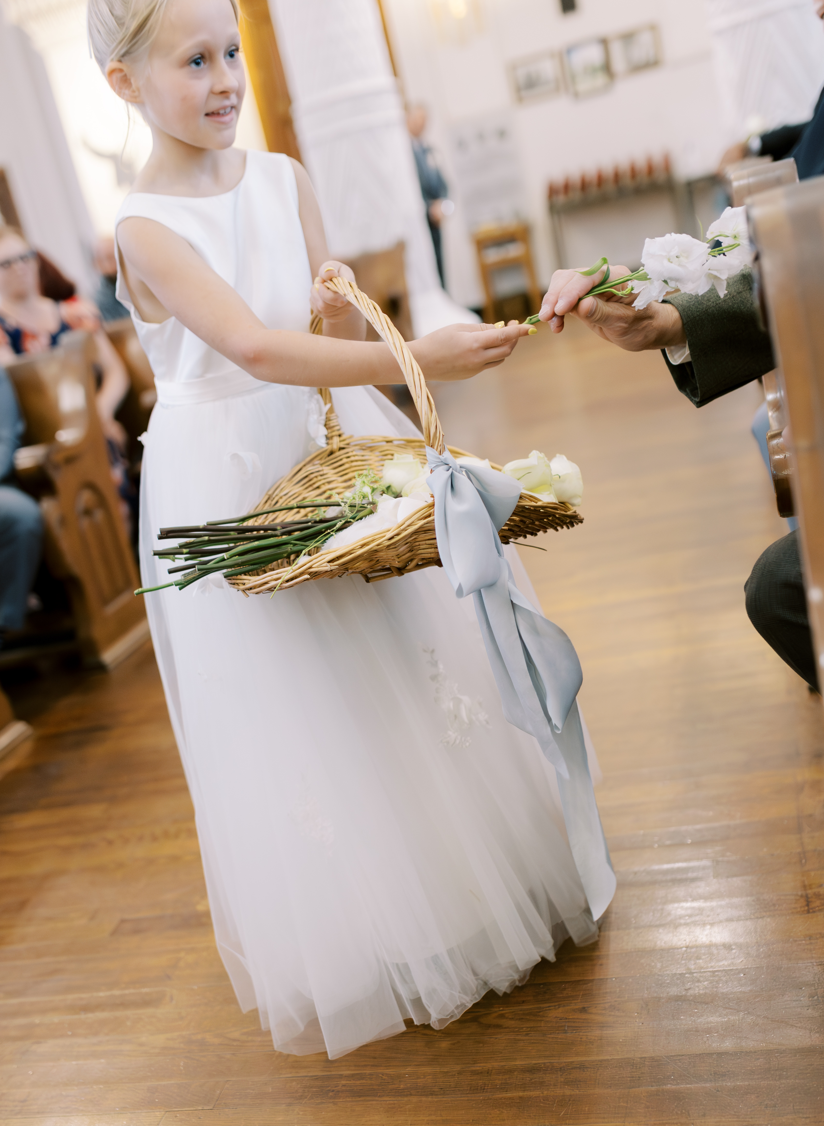 A flower girl in a white dress carries a woven basket of white roses tied with a soft blue ribbon.