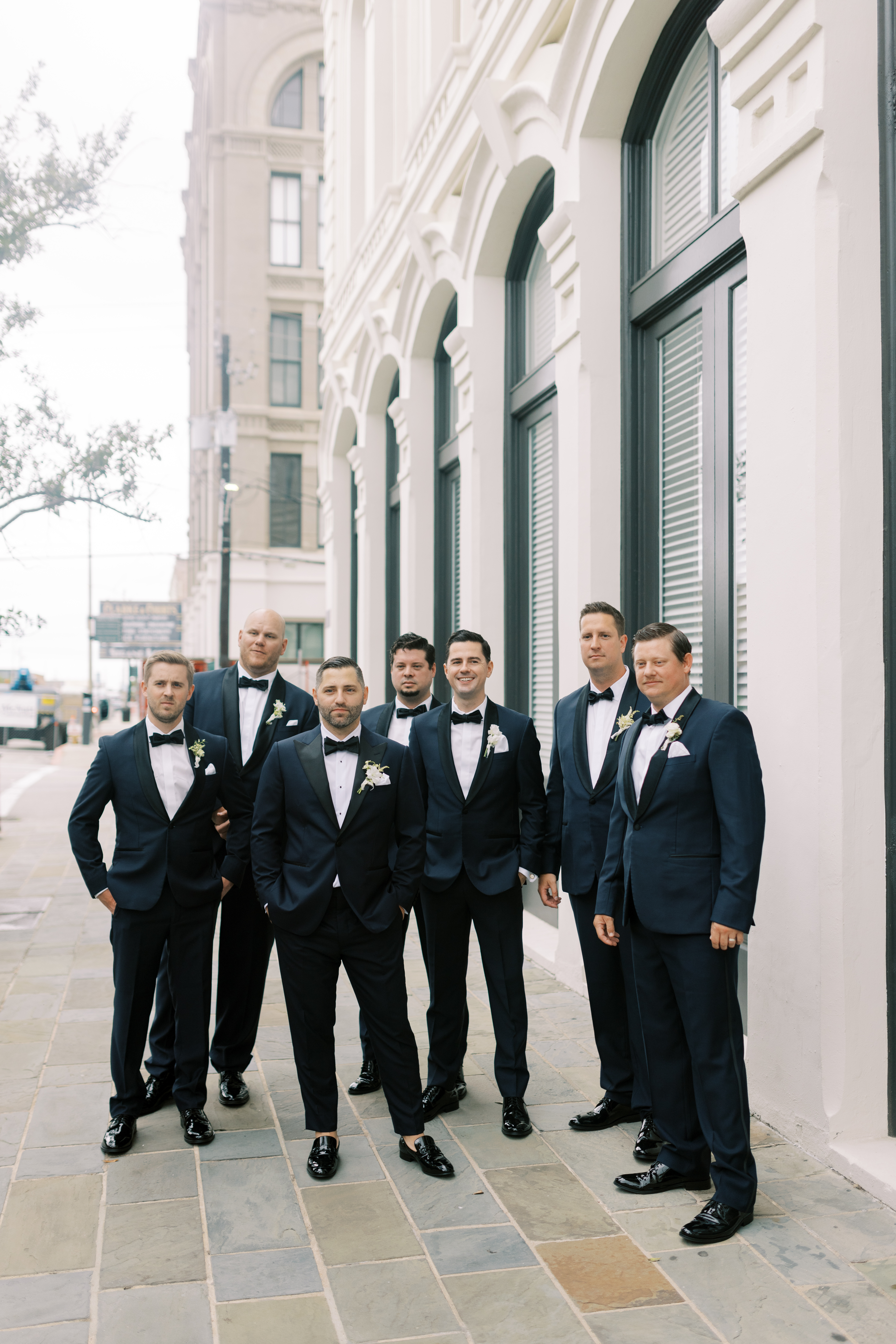 A smiling groom and groomsmen in dark tuxedos with white boutonnieres stand alongside a classical white arched facade.
