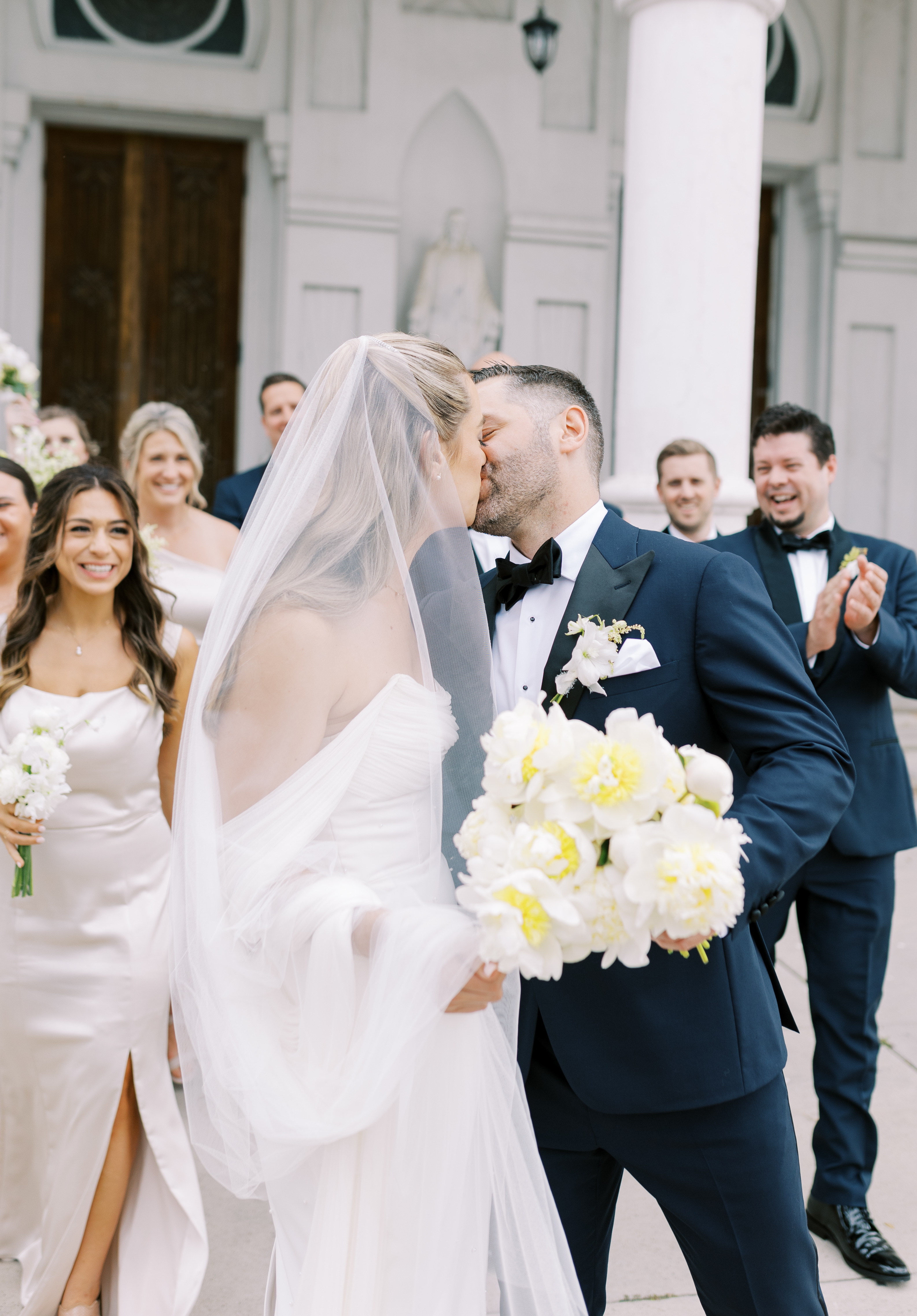 A bride with a tulle veil and white and yellow bouquet kisses a groom in a classic tuxedo outside an arched building.