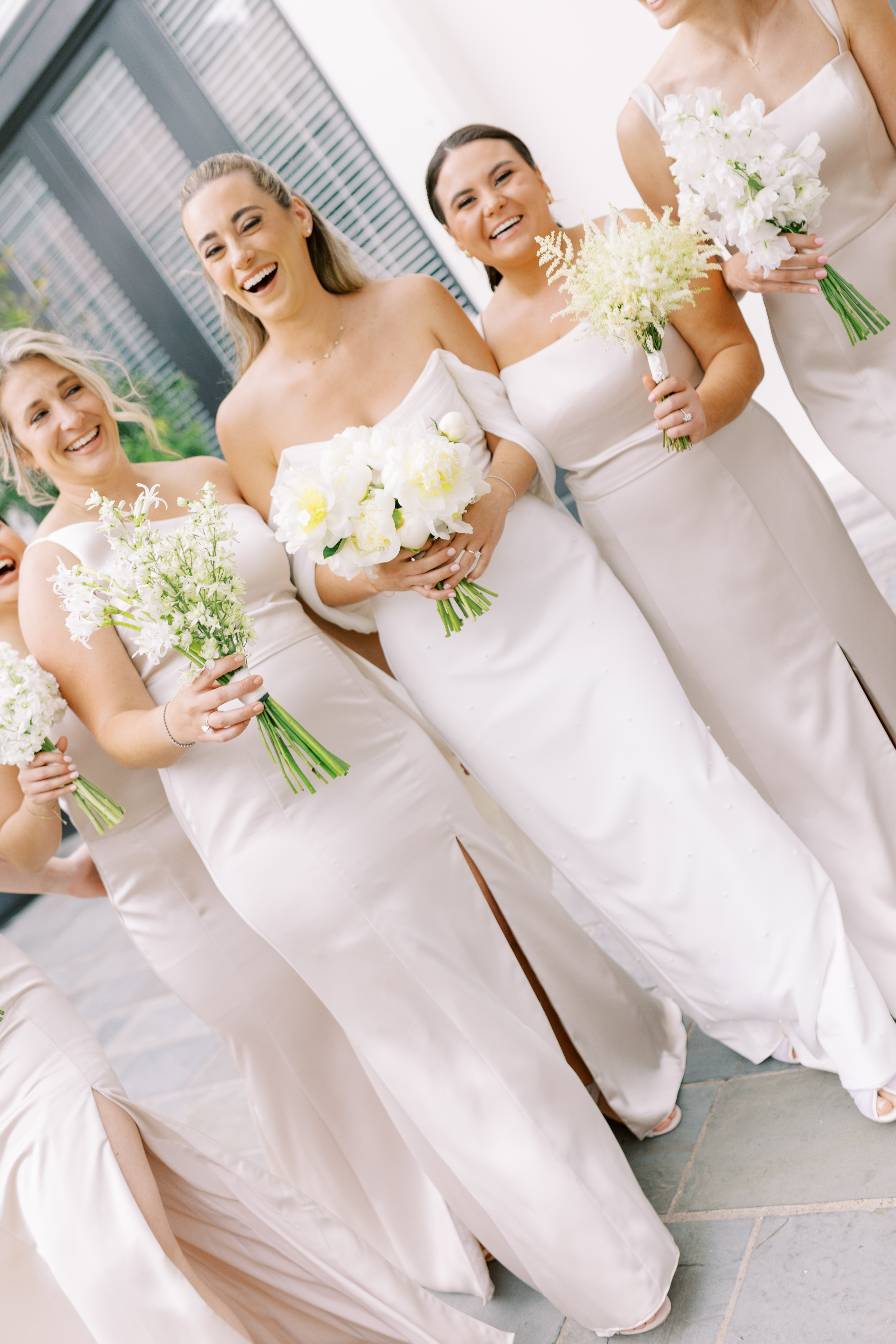 A bride in an off-the-shoulder gown holds a lush bouquet of white peonies beside her bridesmaid.