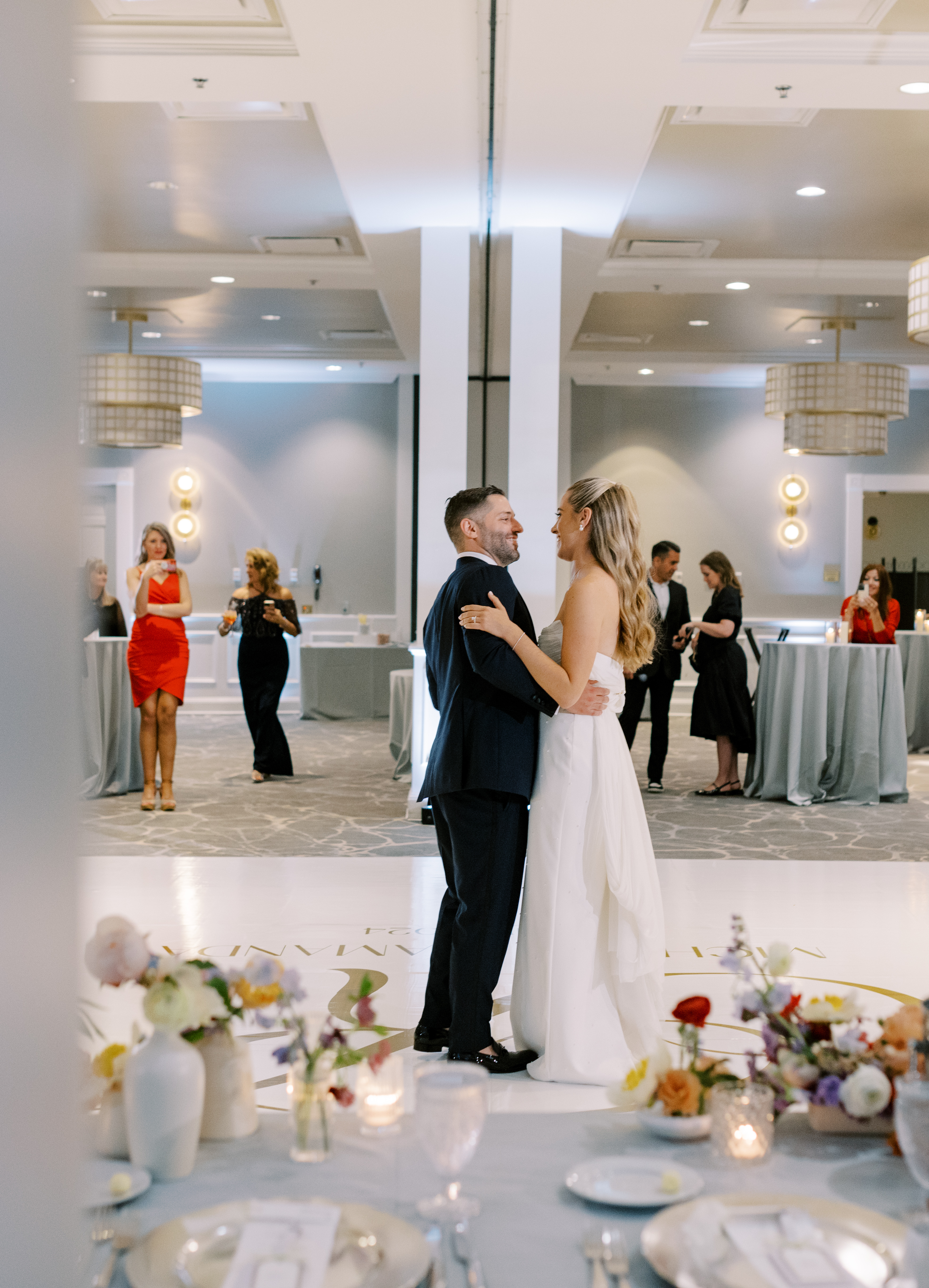 A bride and groom smile at each other while sharing their first dance in an elegant ballroom.