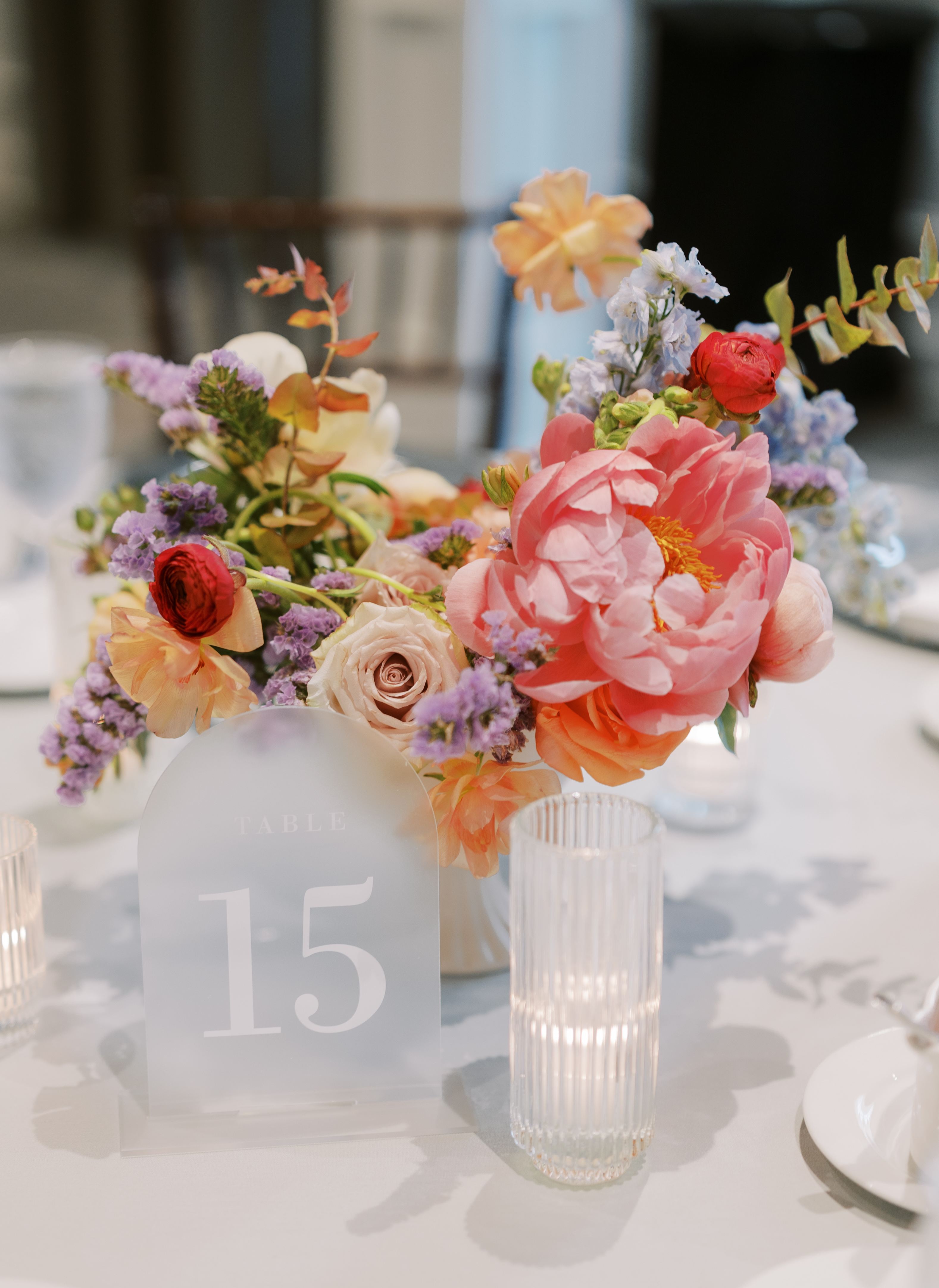A wedding table centerpiece with coral peonies, blush roses, a frosted table number, and a glowing fluted candle.