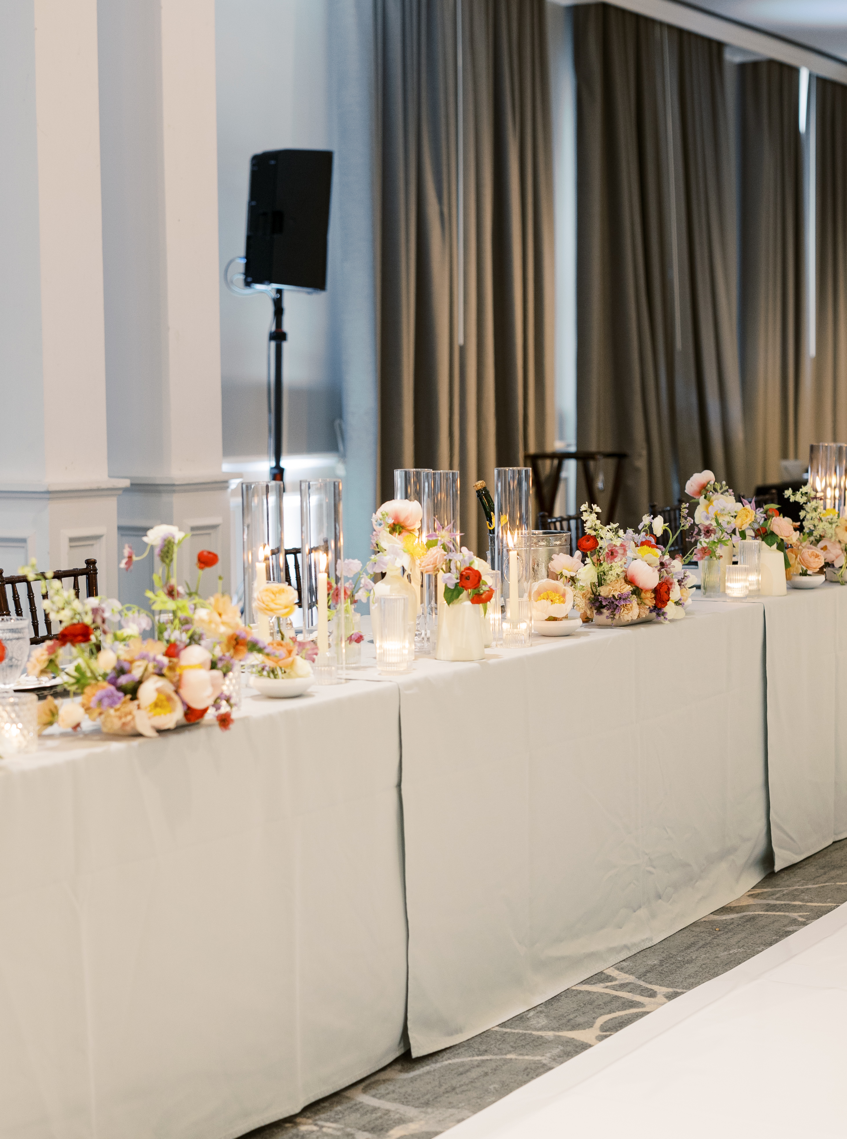 Reception table decorated with white drapery, lit taper candles in glass hurricane vases, and delicate pastel floral arrangements.
