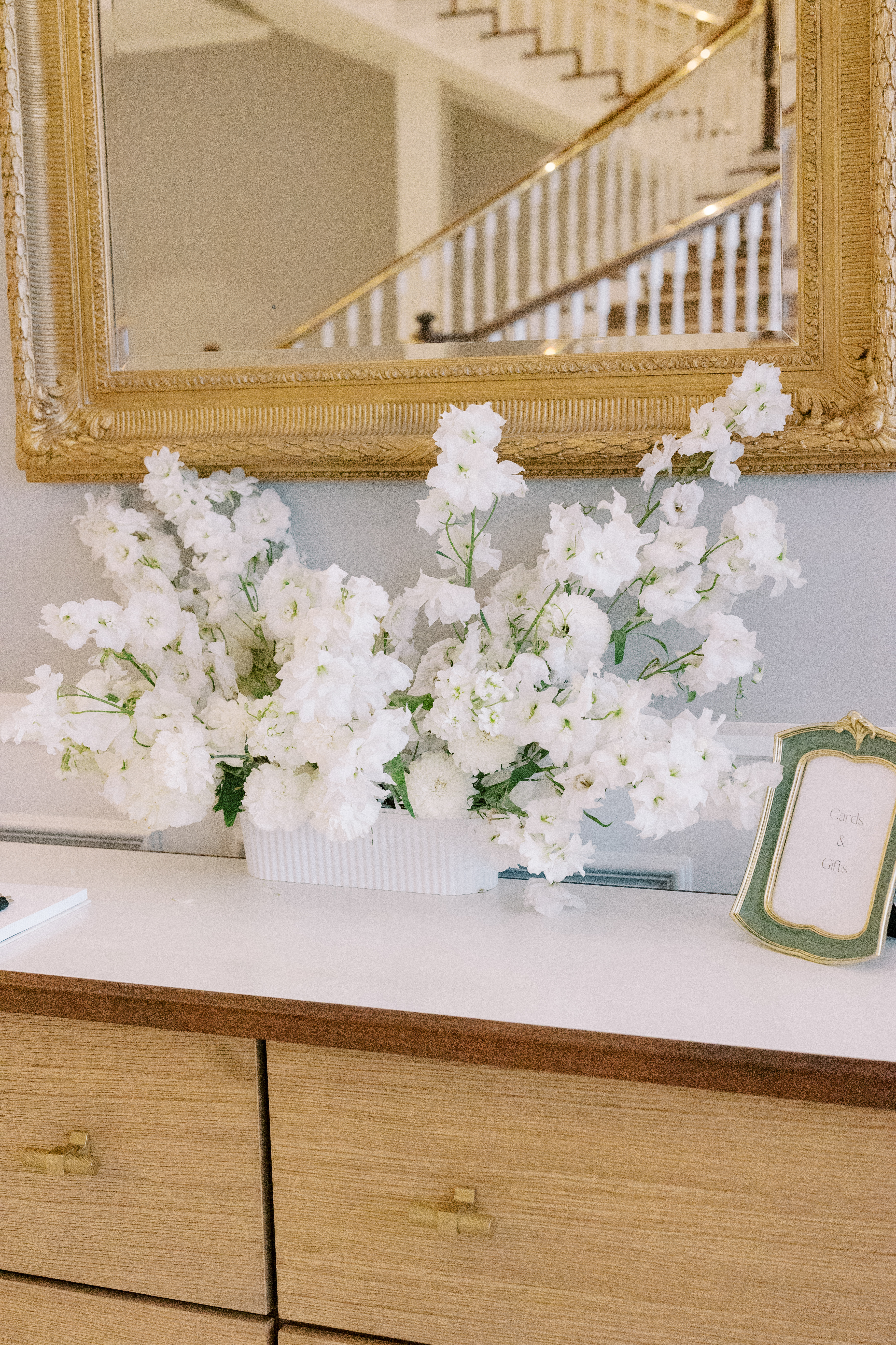 A lush arrangement of white delphiniums and floral blooms sits on a wooden console in front of an ornate gilded mirror.