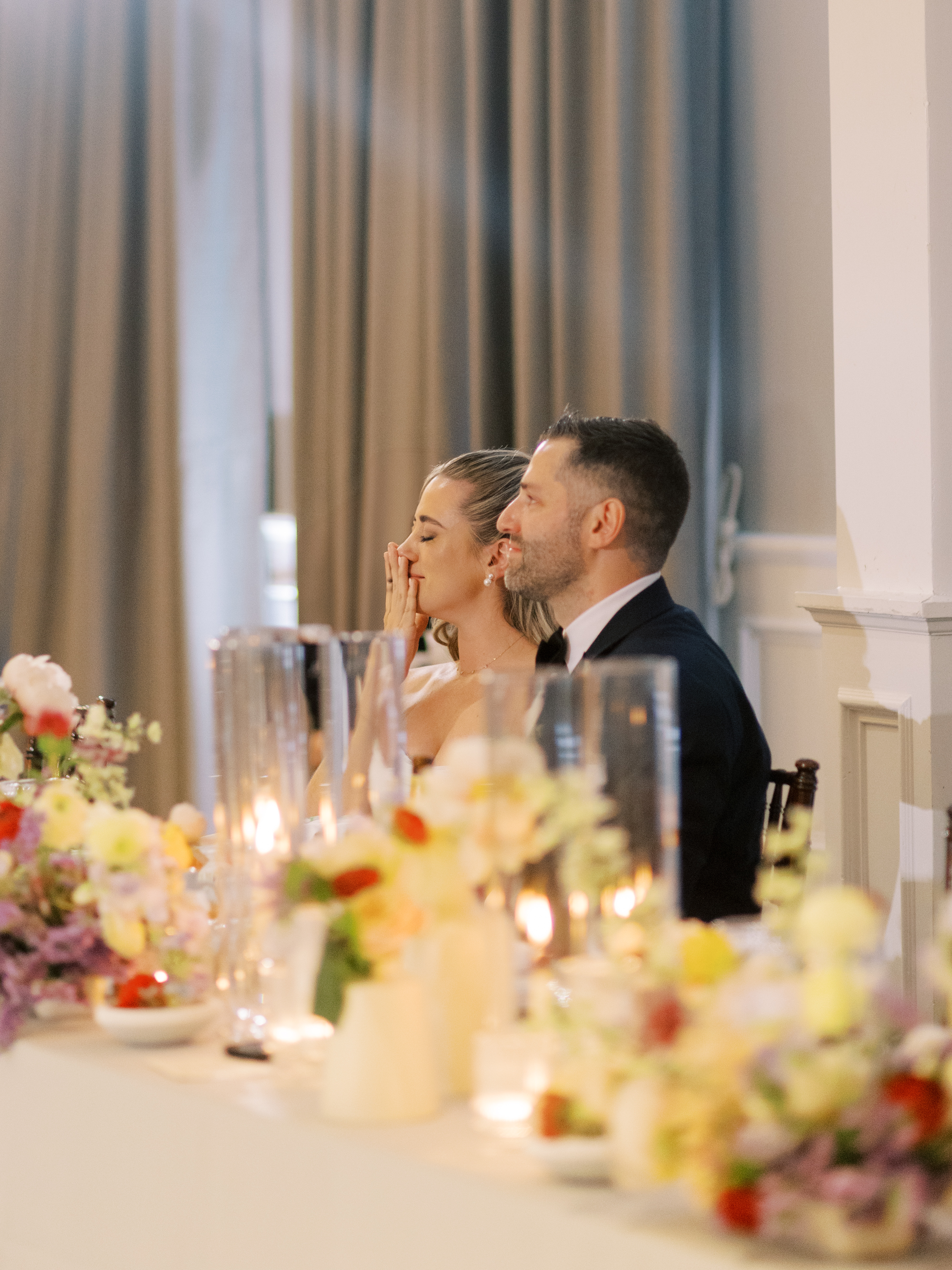 Bride and groom smiling during reception speeches seated behind candlelit tables with floral arrangements.