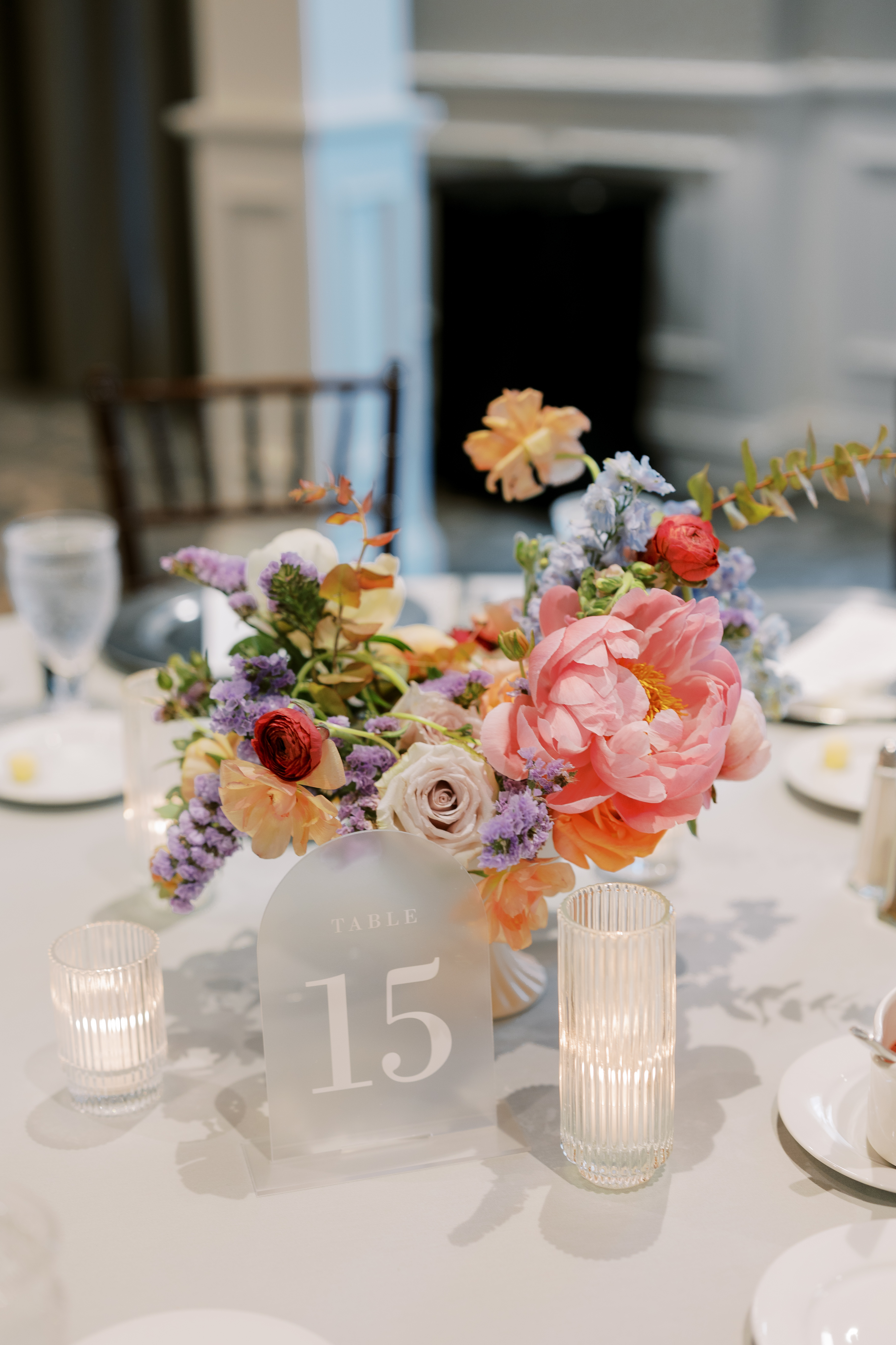 Frosted acrylic table number beside a vibrant floral centerpiece with roses, statice, and a ribbed glass candleholder.