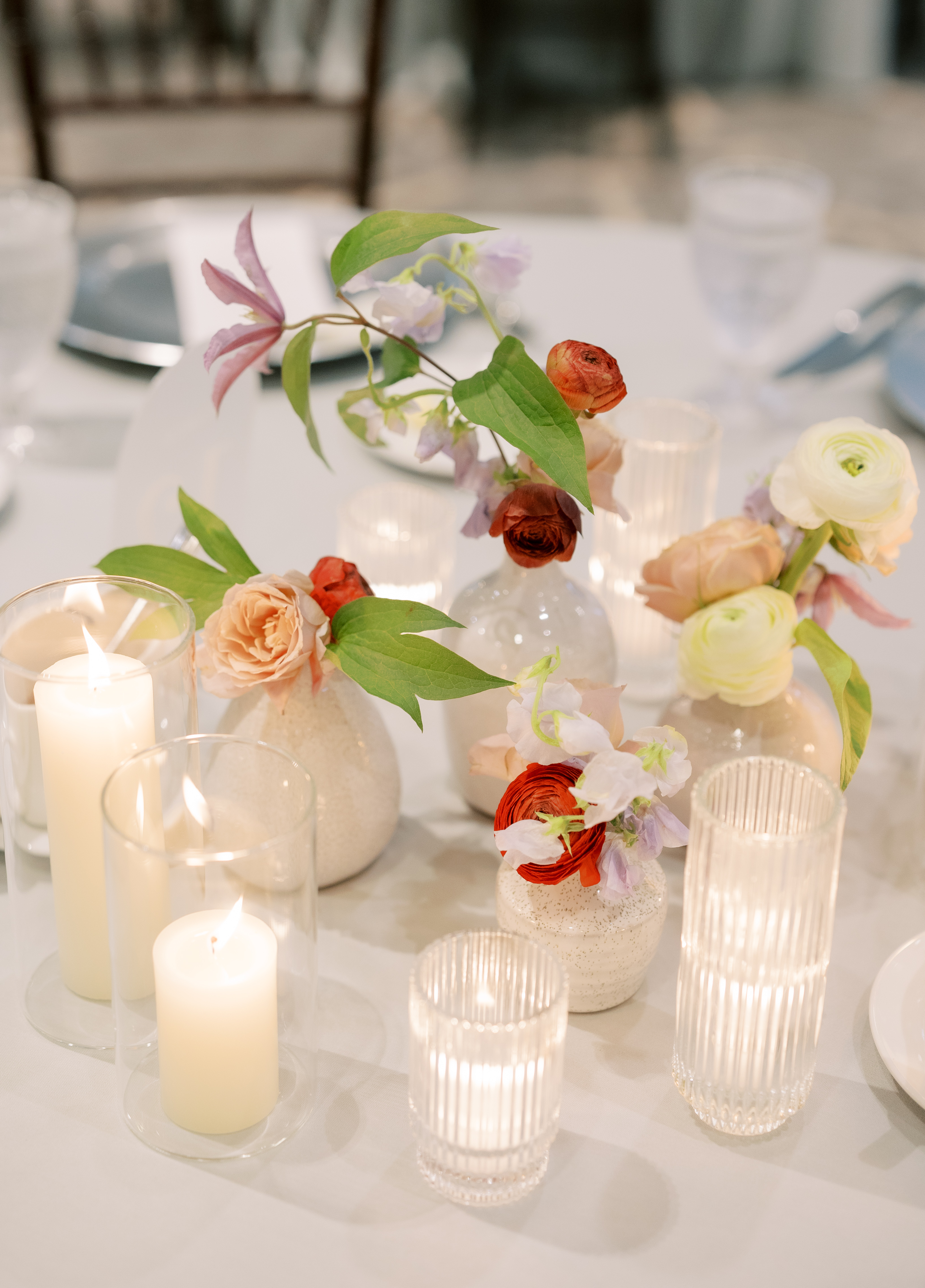 Ceramic bud vases with deep red ranunculus and delicate sweet peas sit alongside fluted glass candles on an elegant reception table.