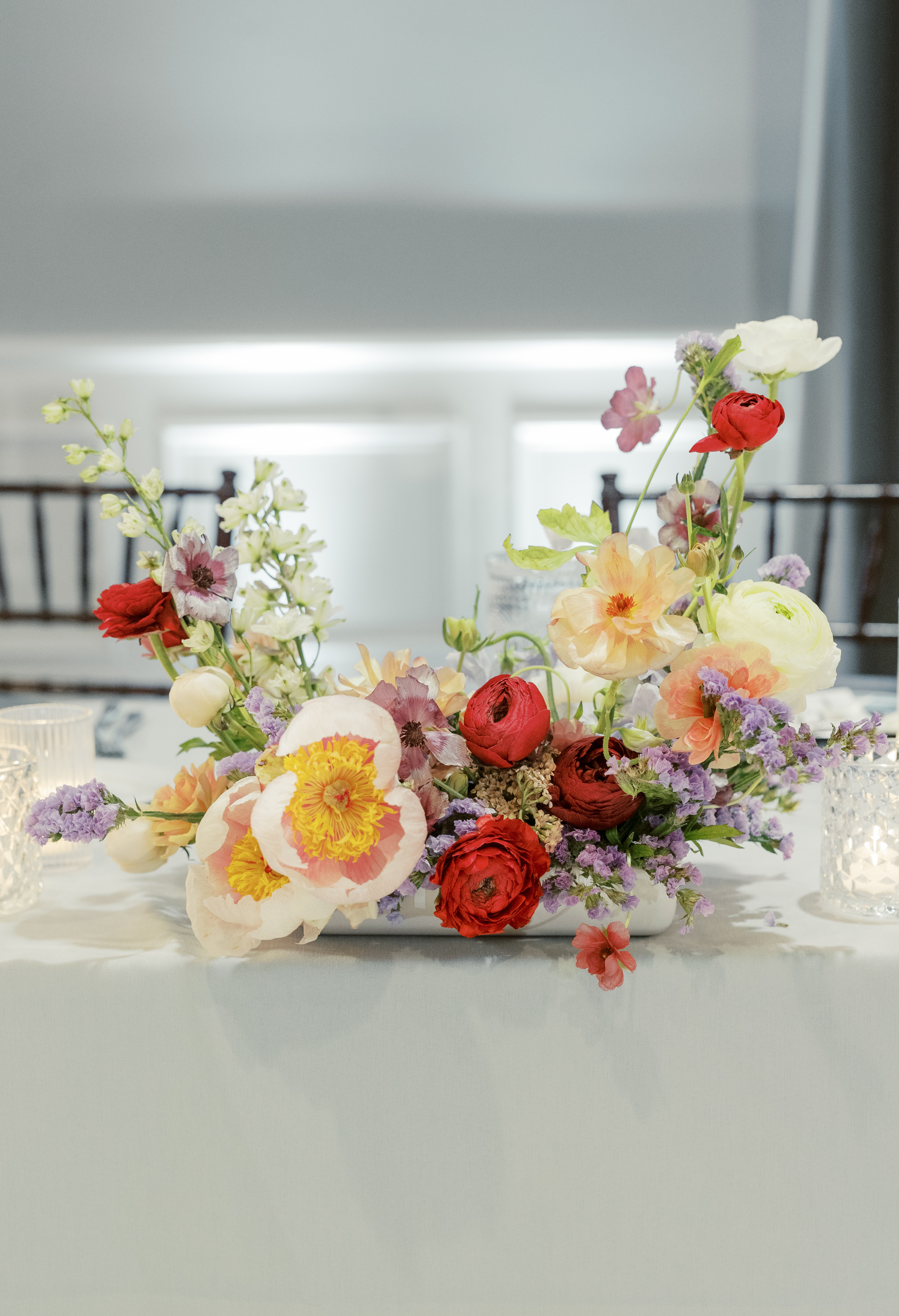 A colorful floral arrangement featuring deep red ranunculus, an open blush poppy, and delicate pastel blooms on a tabletop.