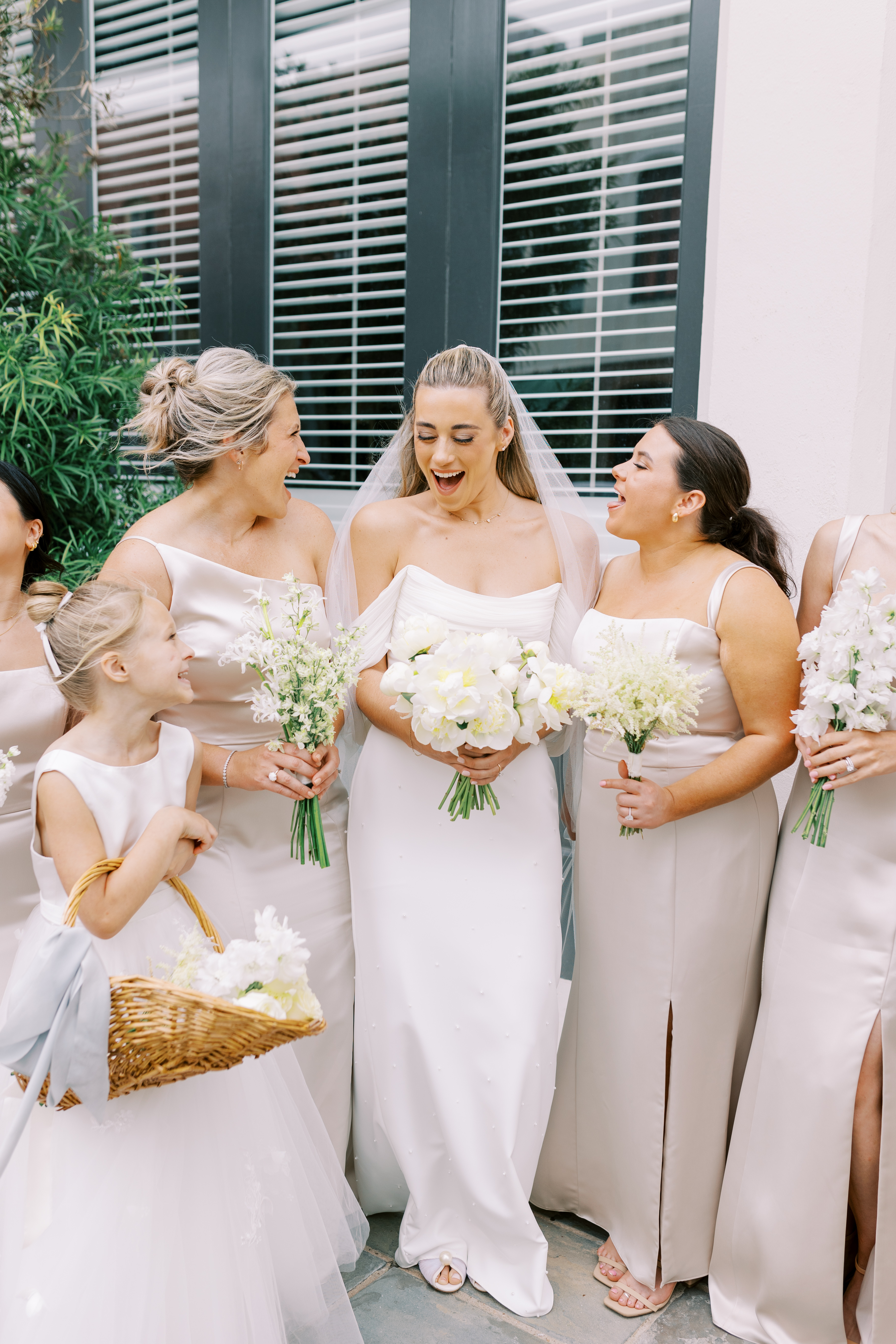 A laughing bride in an off-the-shoulder gown holds an all-white peony bouquet alongside her bridesmaids.