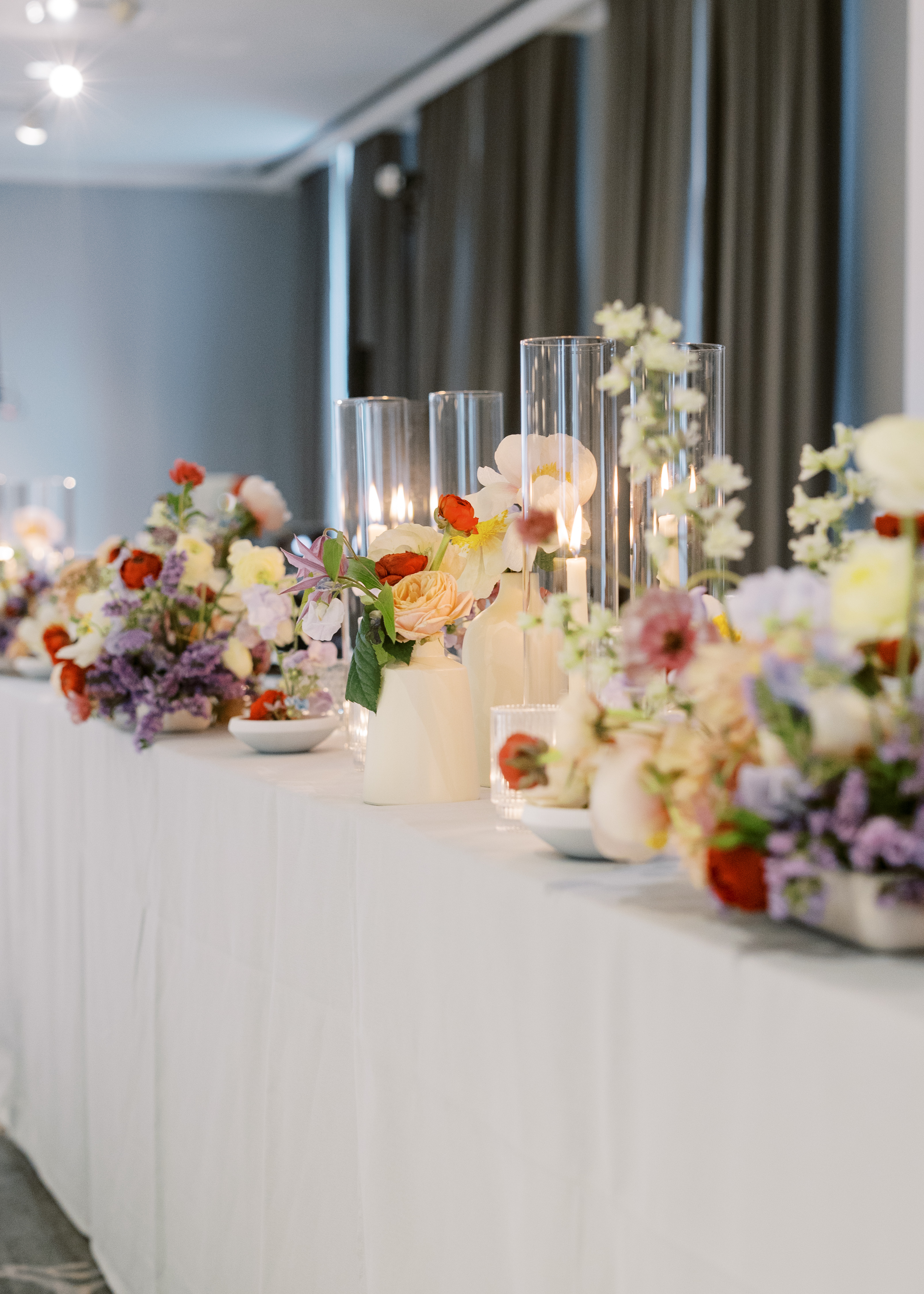 Delicate floral bud vases and glowing glass taper candles line a white linen reception table.
