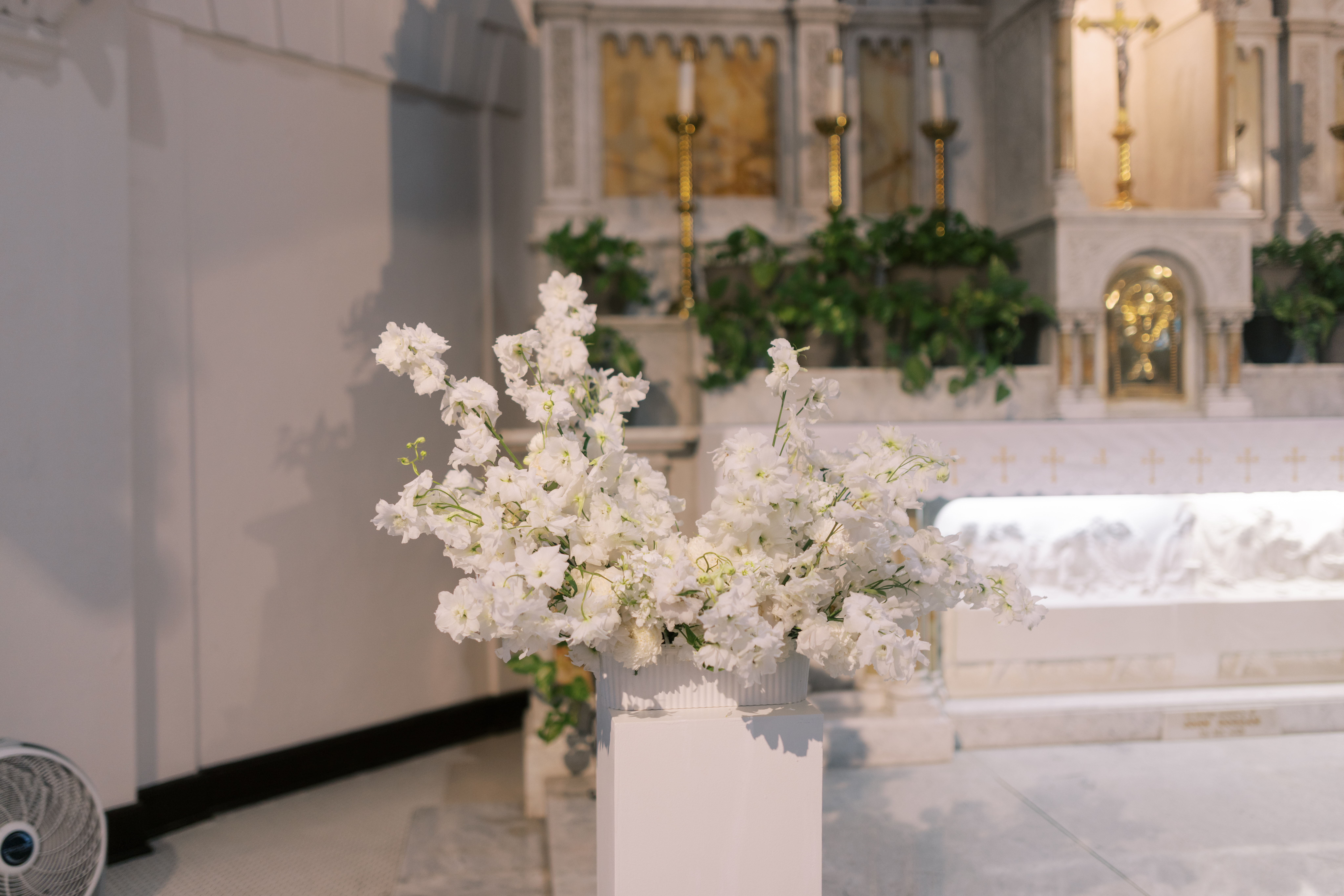Lush white delphiniums and floral arrangements in a fluted vase set on a white pedestal against a warm stone backdrop.