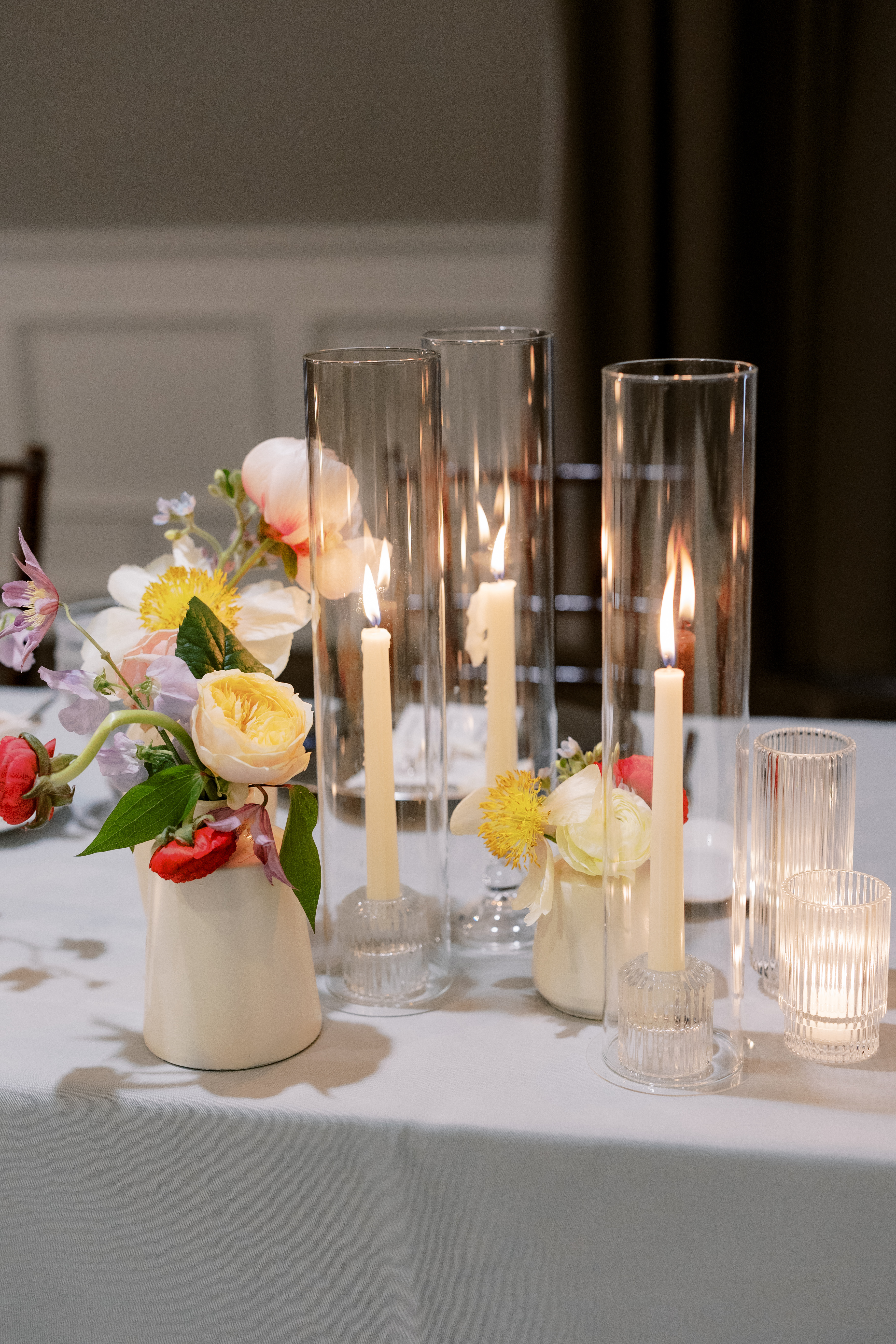 Lit taper candles in tall clear glass hurricane sleeves set among soft yellow and white flowers on an elegant dining table.