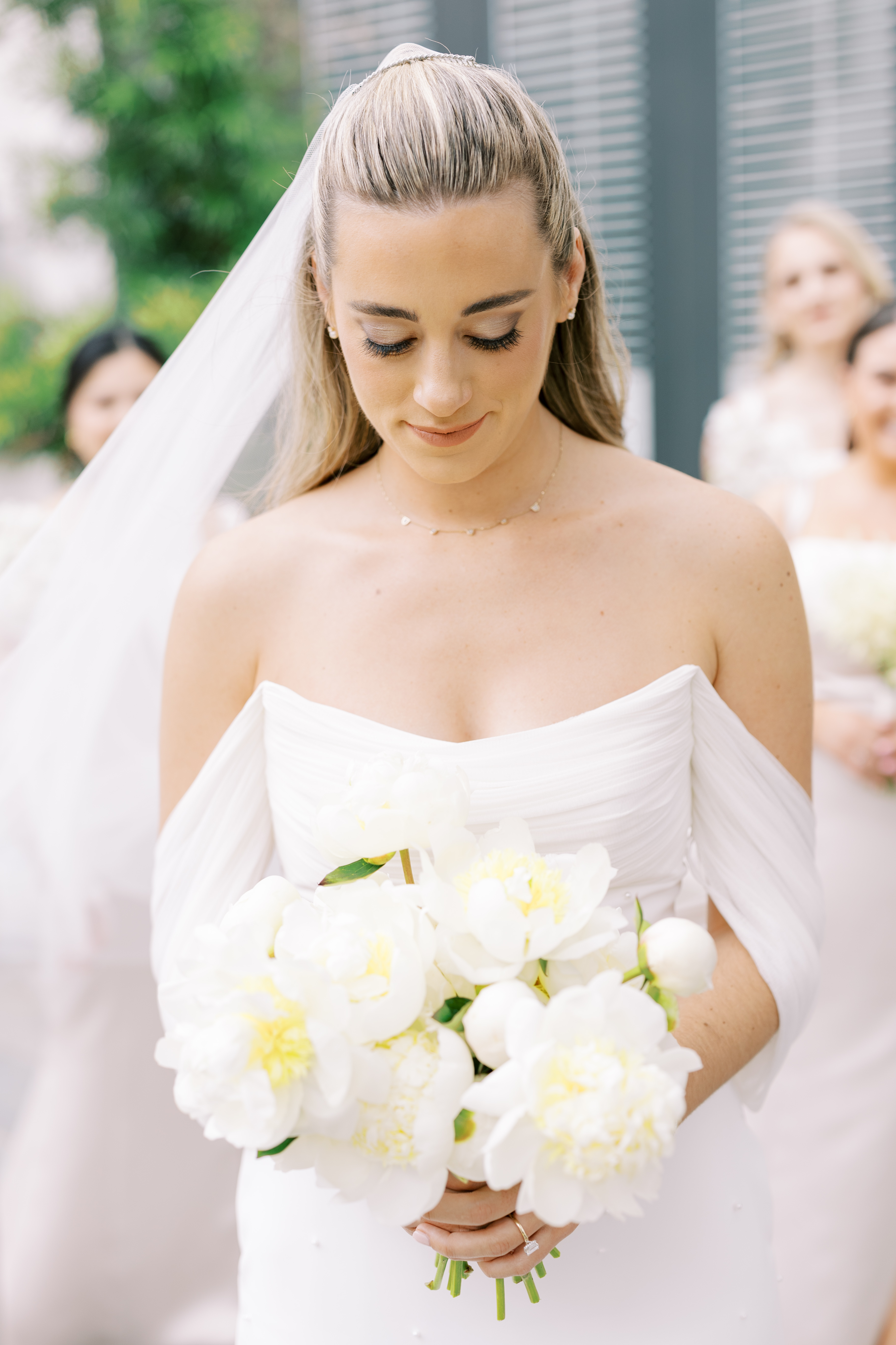 A bride looks softly down at a bouquet of open white peonies with delicate yellow centers.
