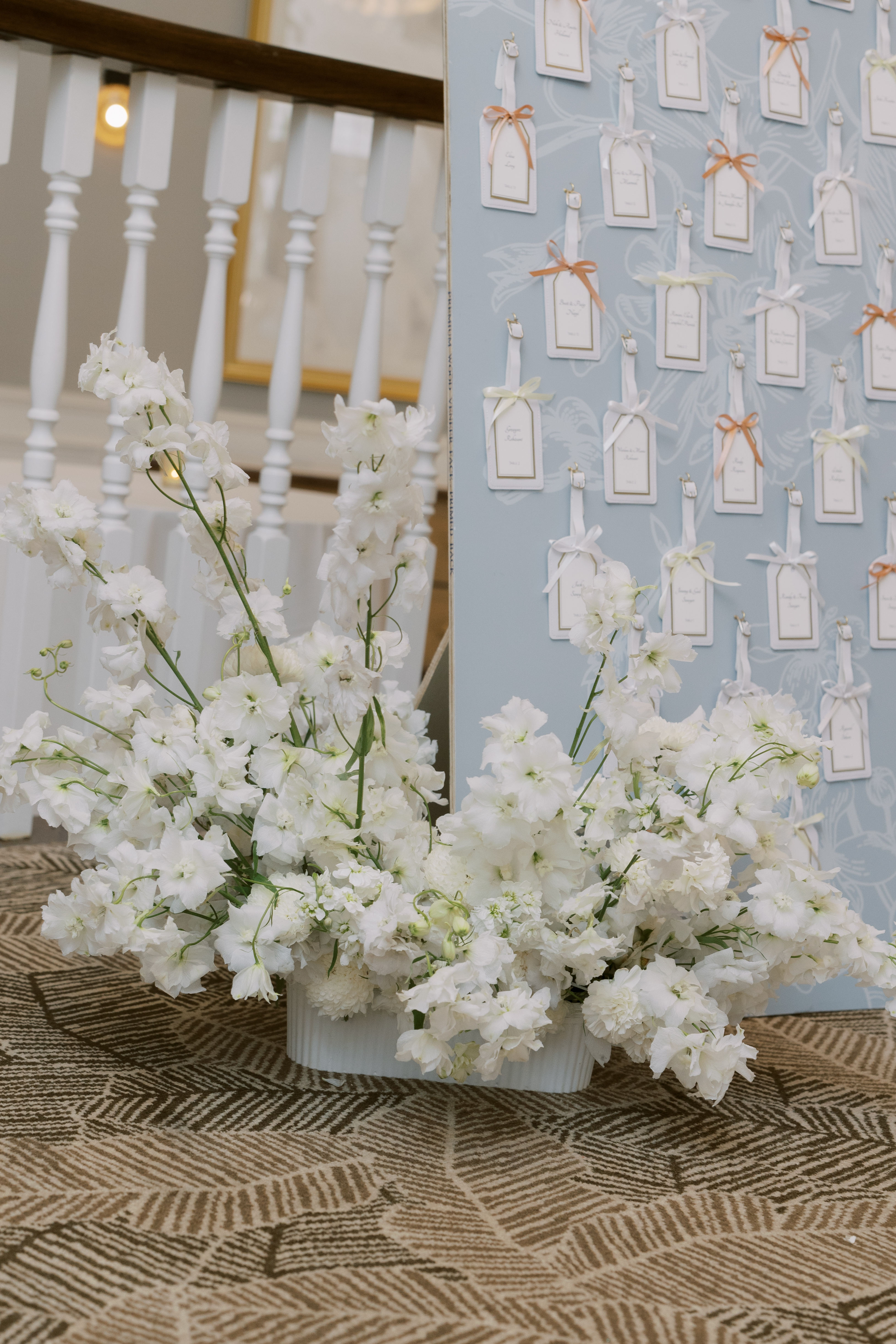White delphiniums and lush florals accent a soft blue seating chart display with ribbon-tied luggage tags.