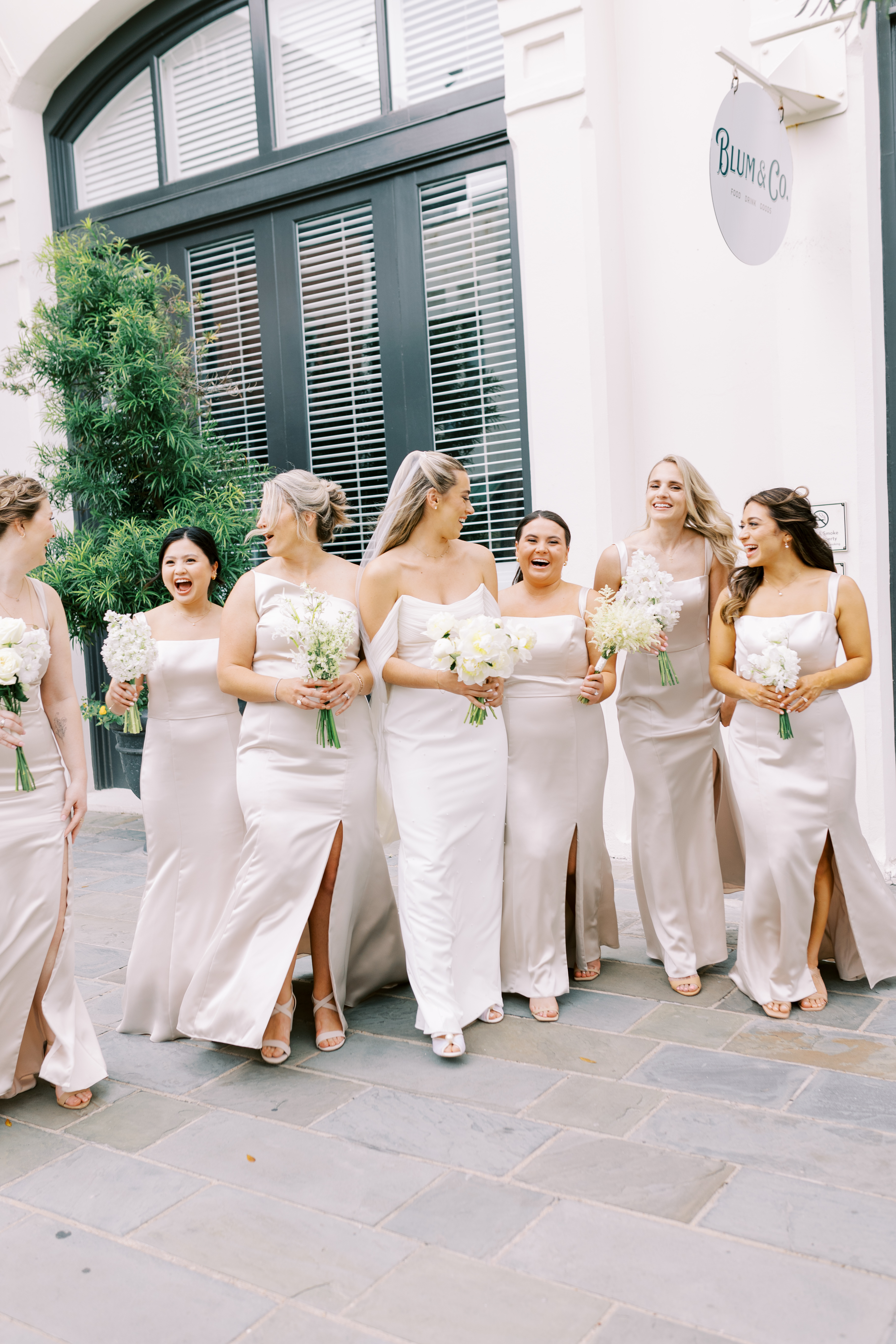 Bride and bridesmaids in elegant neutral gowns smiling together outdoors while carrying delicate white floral bouquets.