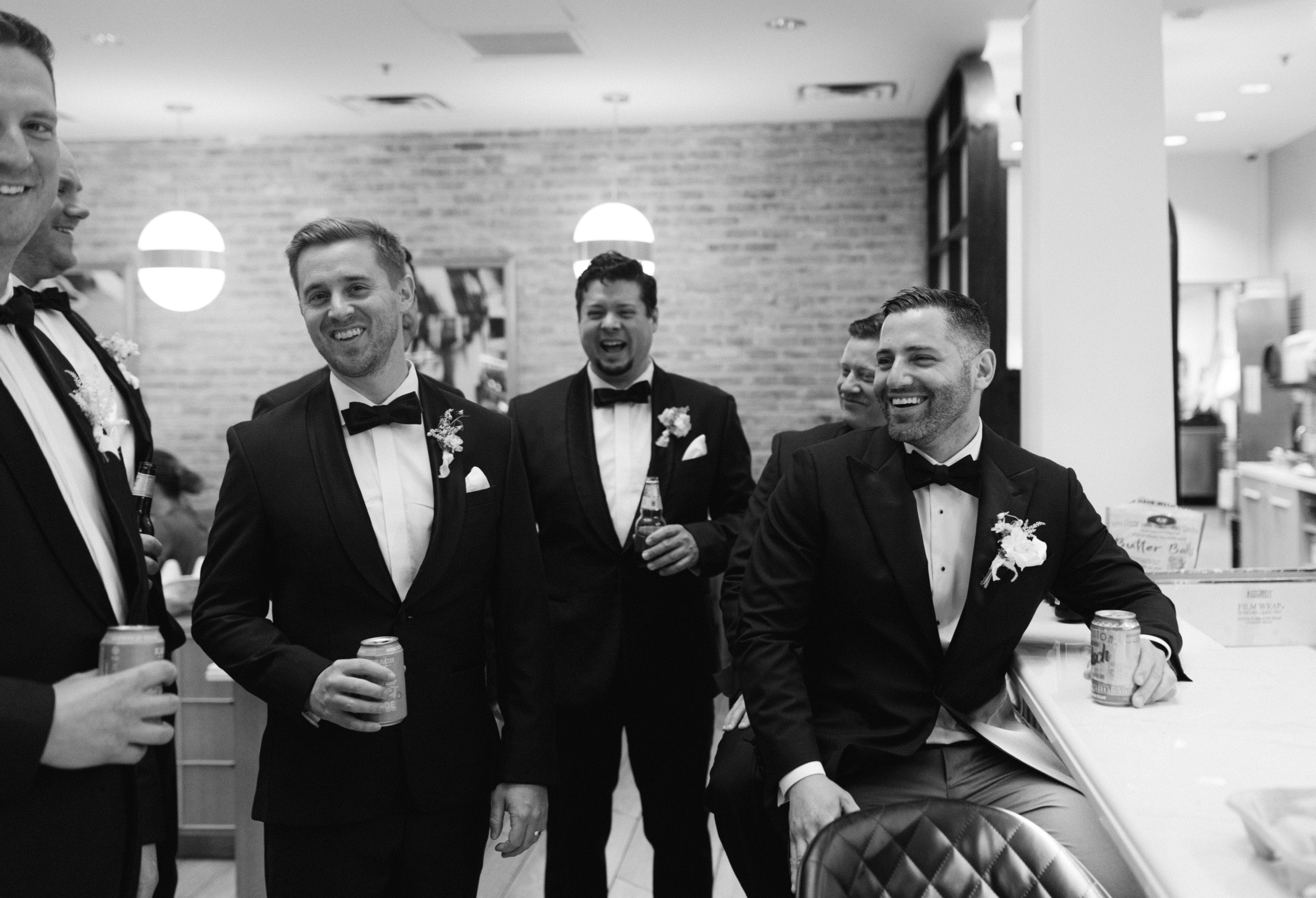A smiling man wearing a black tuxedo with a bow tie and floral boutonnière holds a drink.