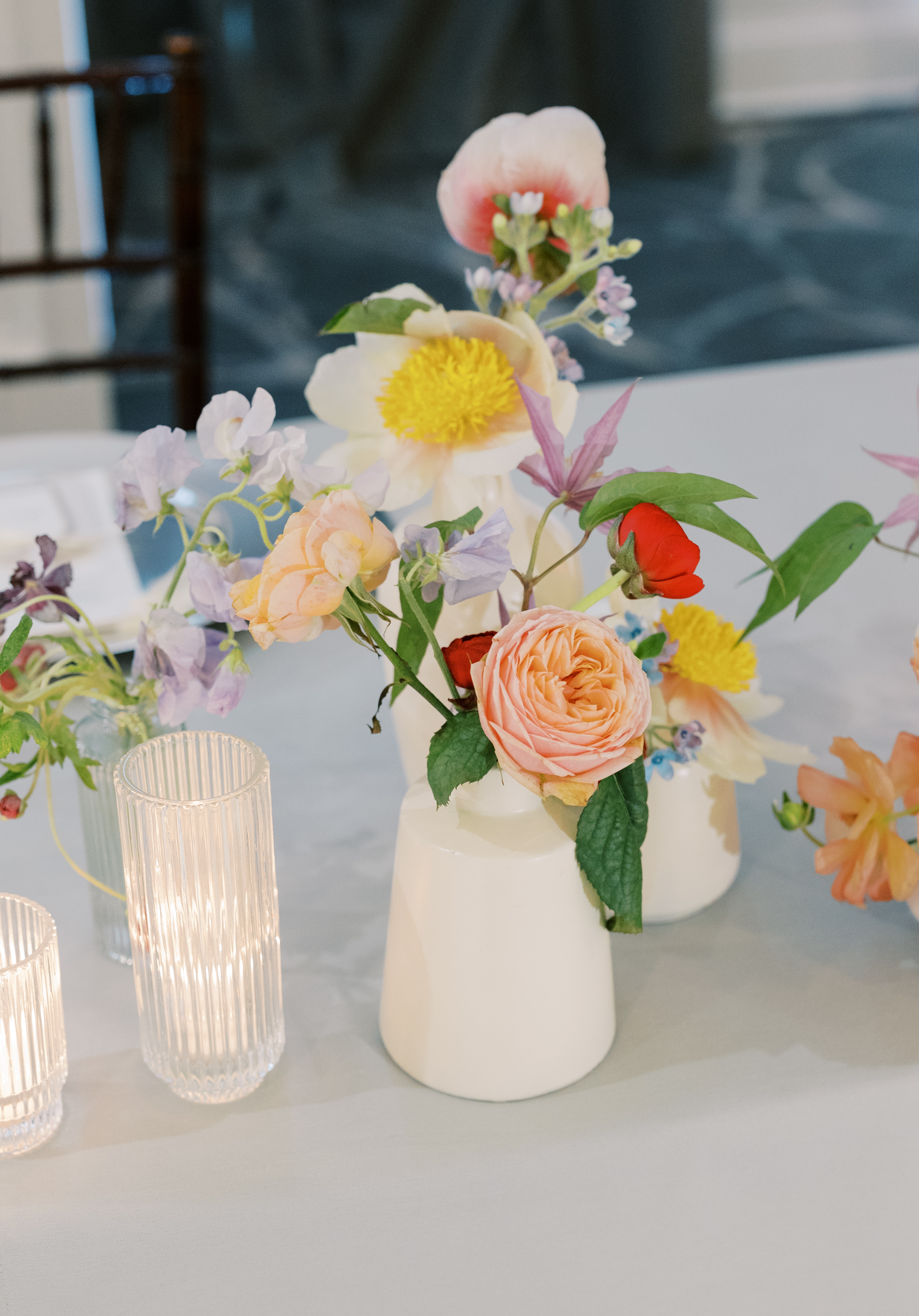 Peach garden roses, poppies, and pastel blooms arranged in white ceramic bud vases on a tabletop.