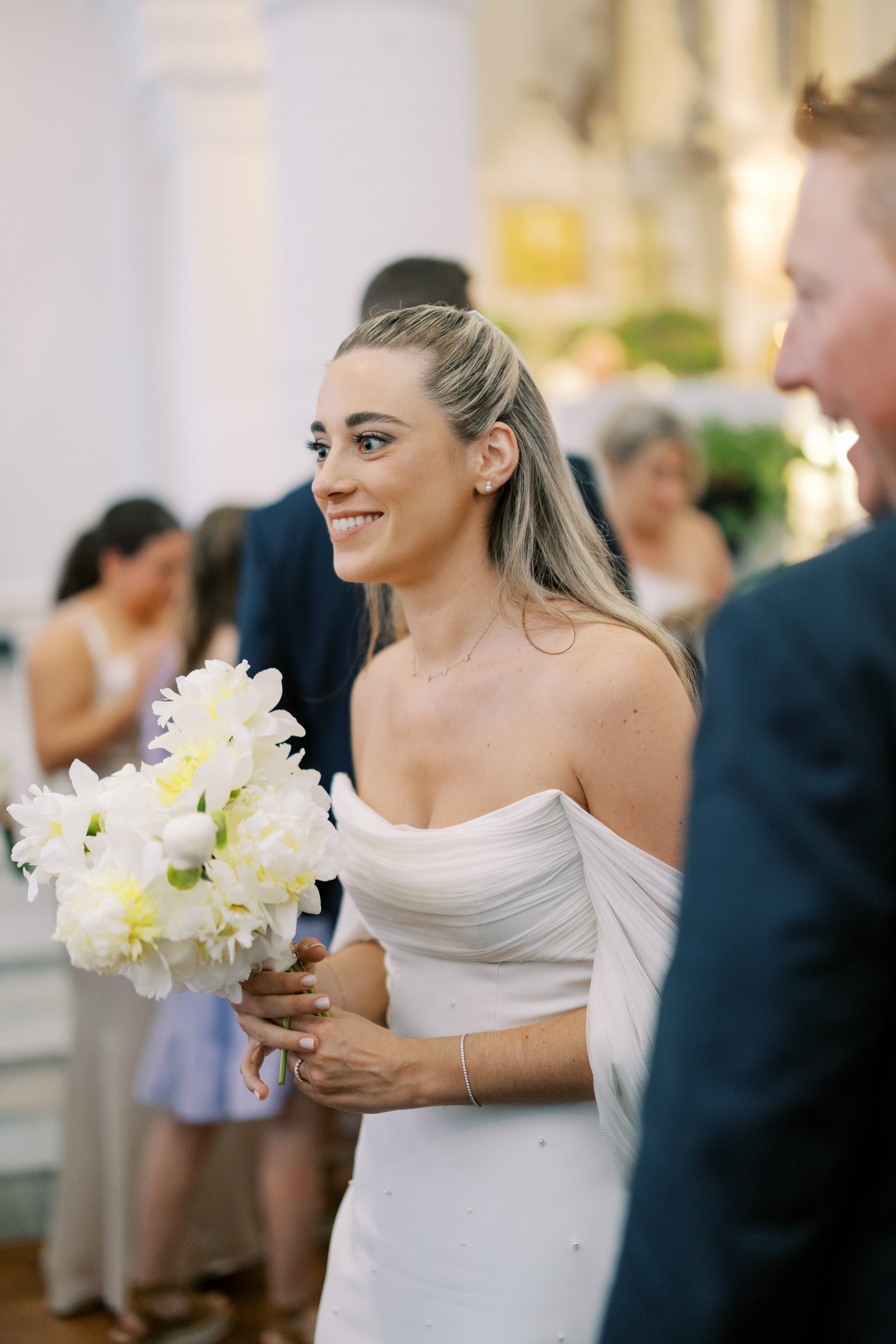Smiling bride in an elegant white gown with a ruched neckline holding a bouquet of white flowers.