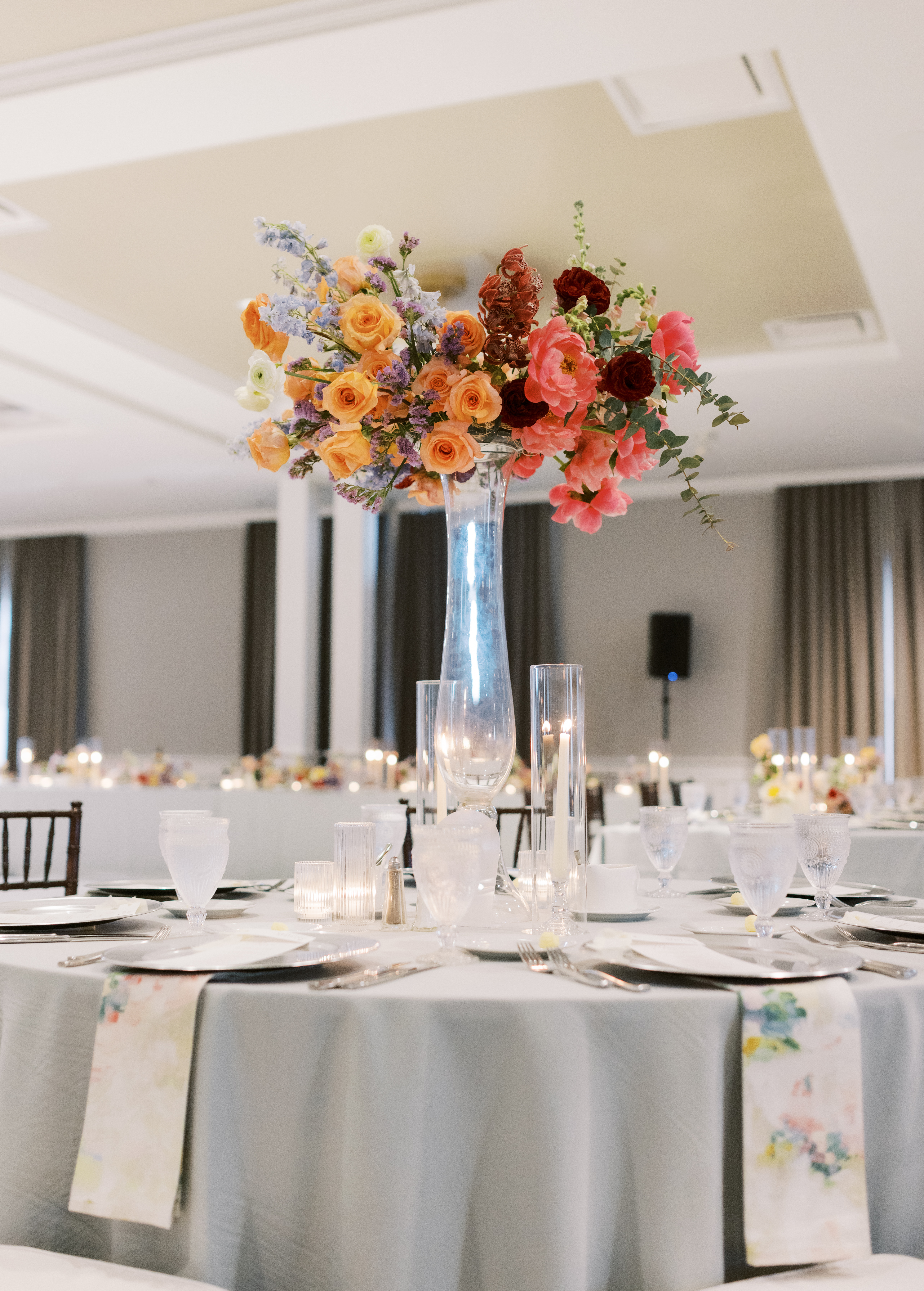 A tall glass vase holds vibrant peach roses, pink peonies, and orchids above an elegant candlelit reception table.