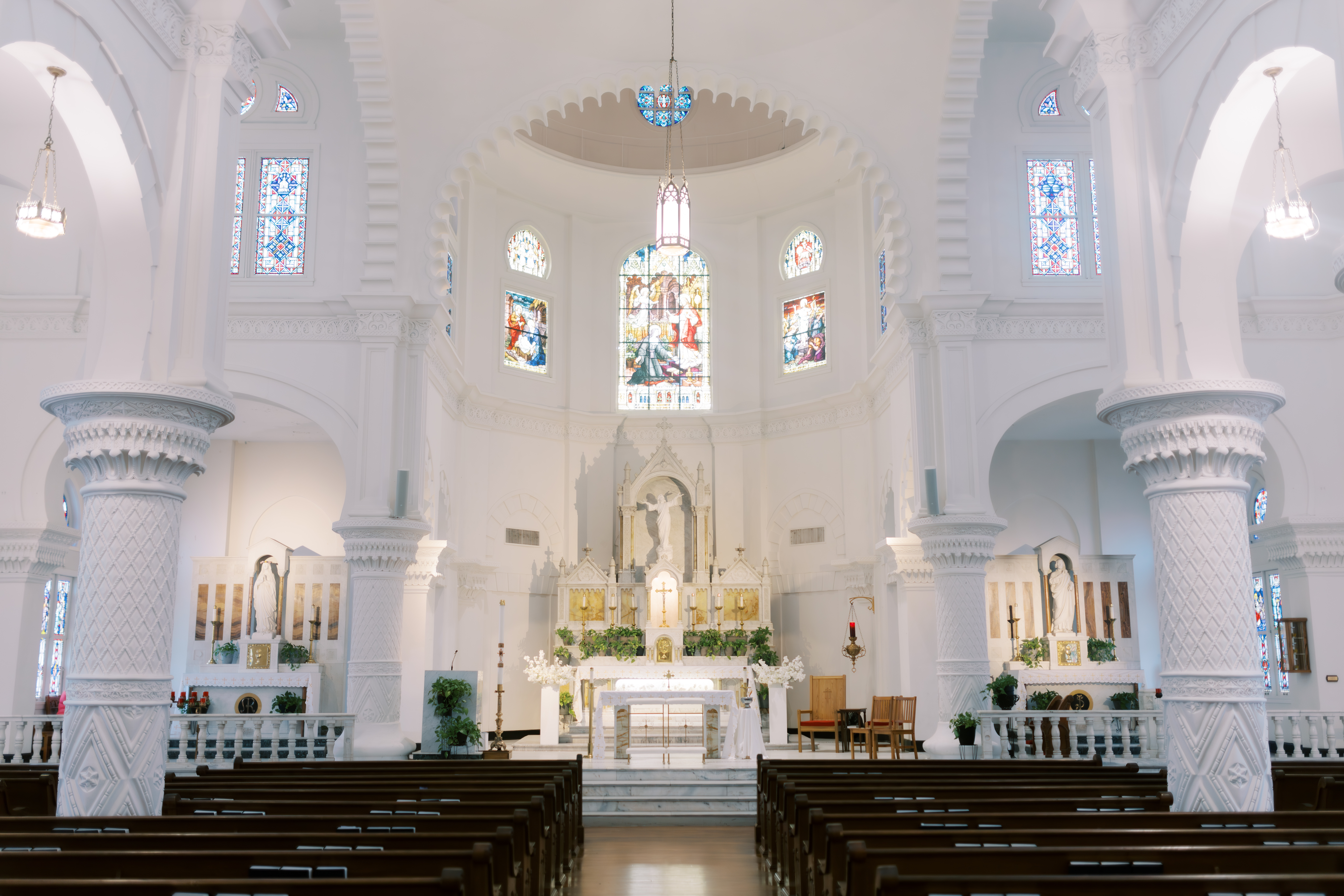 A white church altar with candles, greenery, and kneeling benches sits beneath a tall stained glass window and hanging lantern.