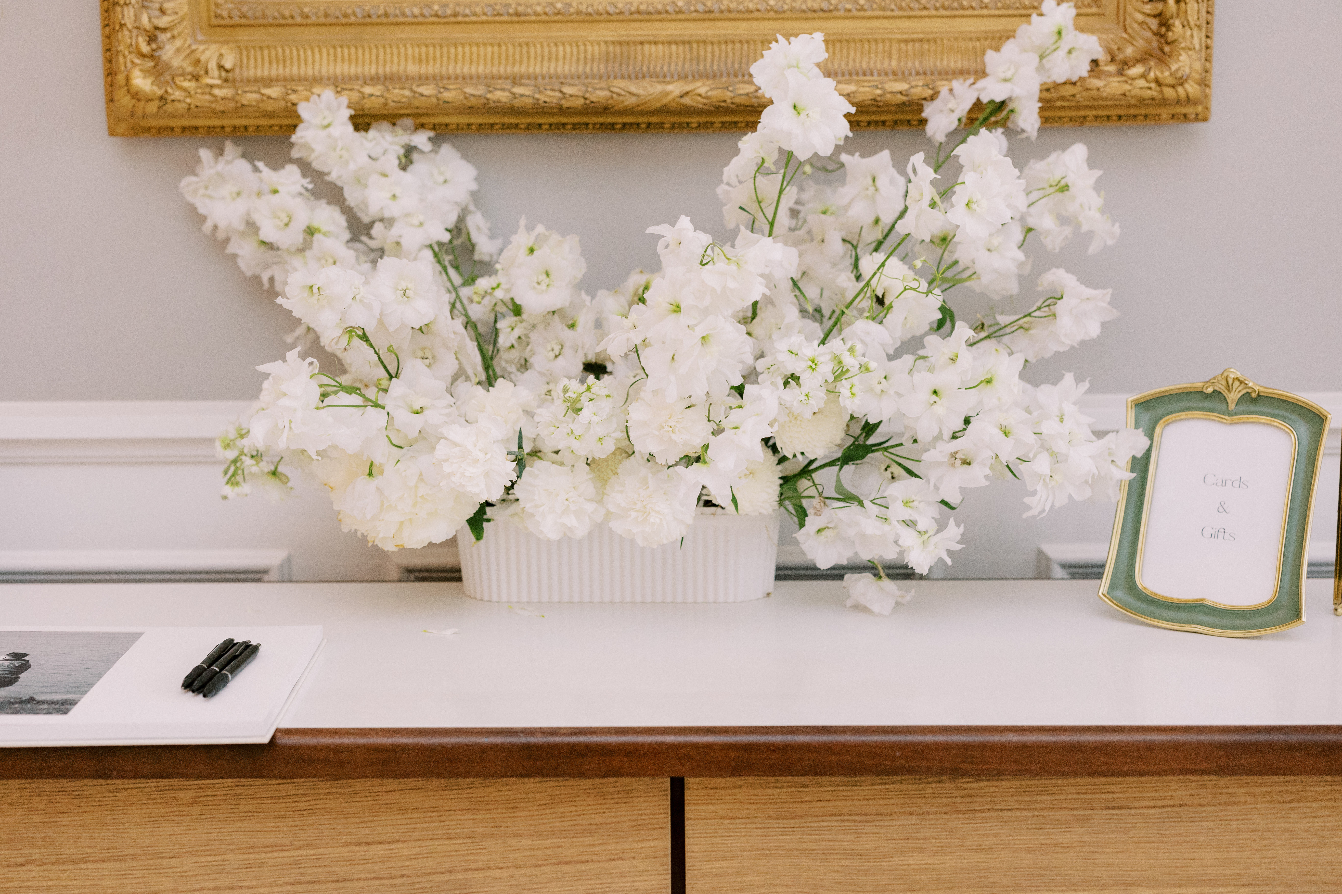 All-white floral arrangement in a fluted container positioned below an ornate gilded frame.