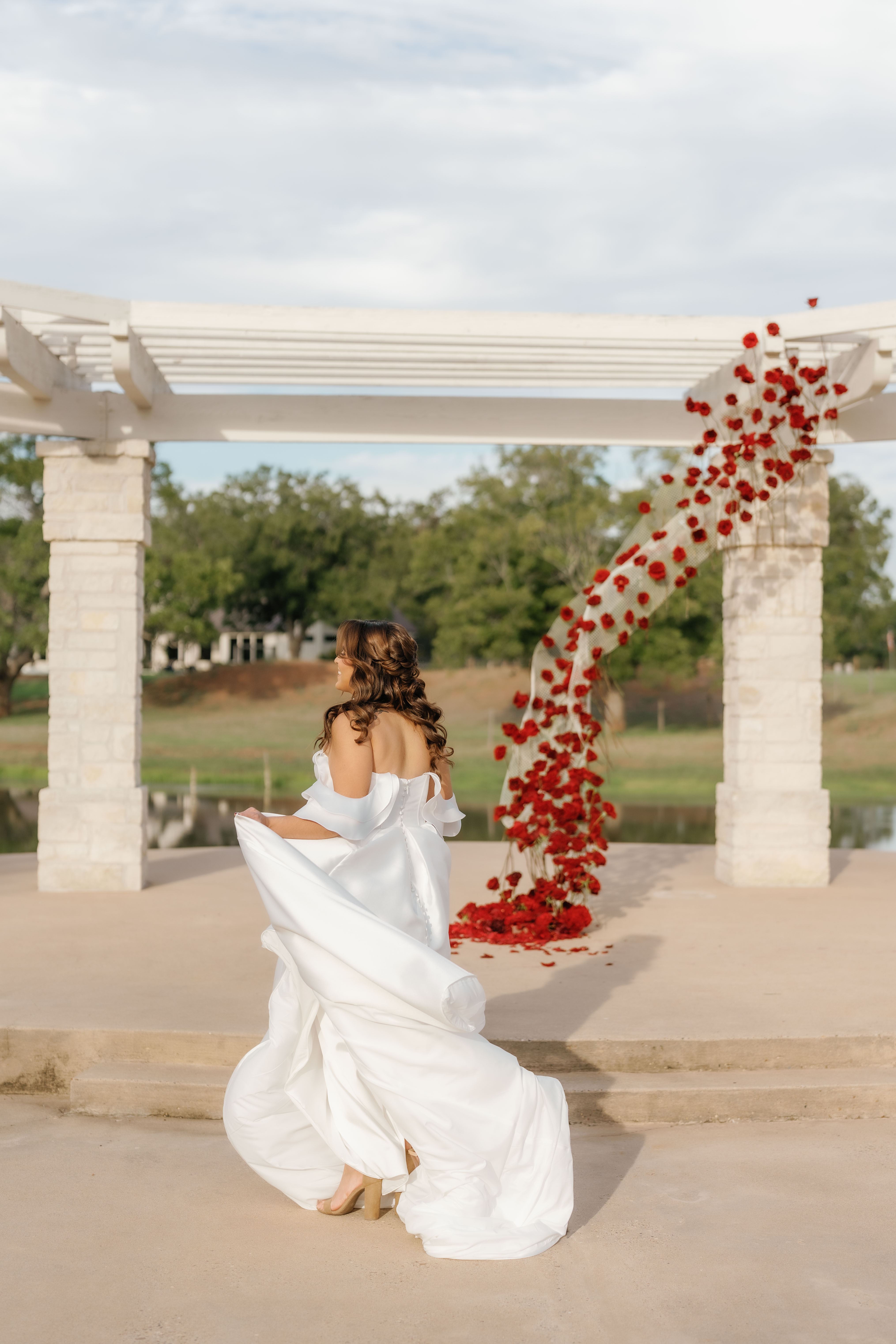 A bride in a white satin gown stands outdoors near a sculptural ceremony backdrop woven with deep red roses.
