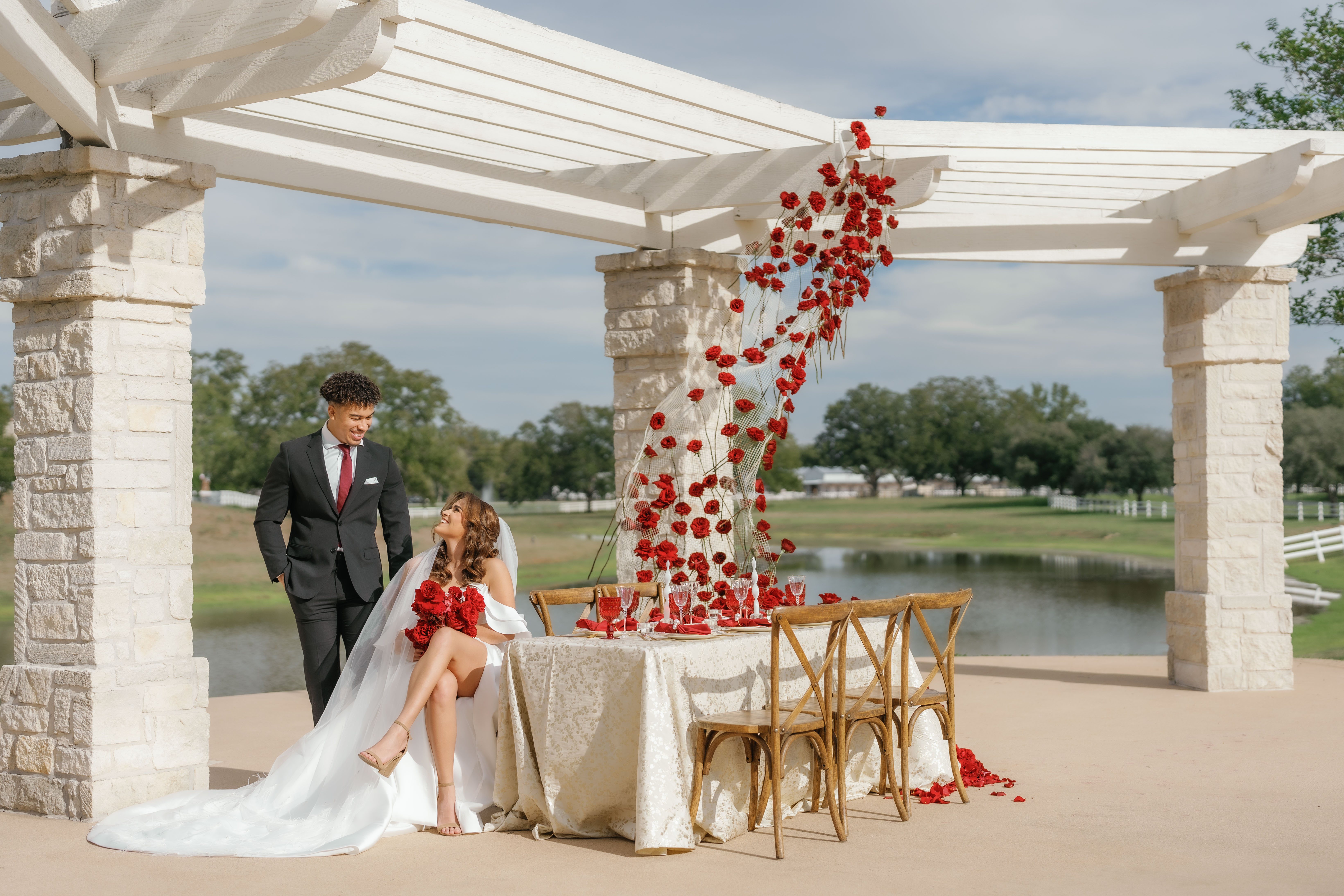 Sculptural red roses suspended on mesh netting rise behind an elegant outdoor reception table set beneath a white pergola.