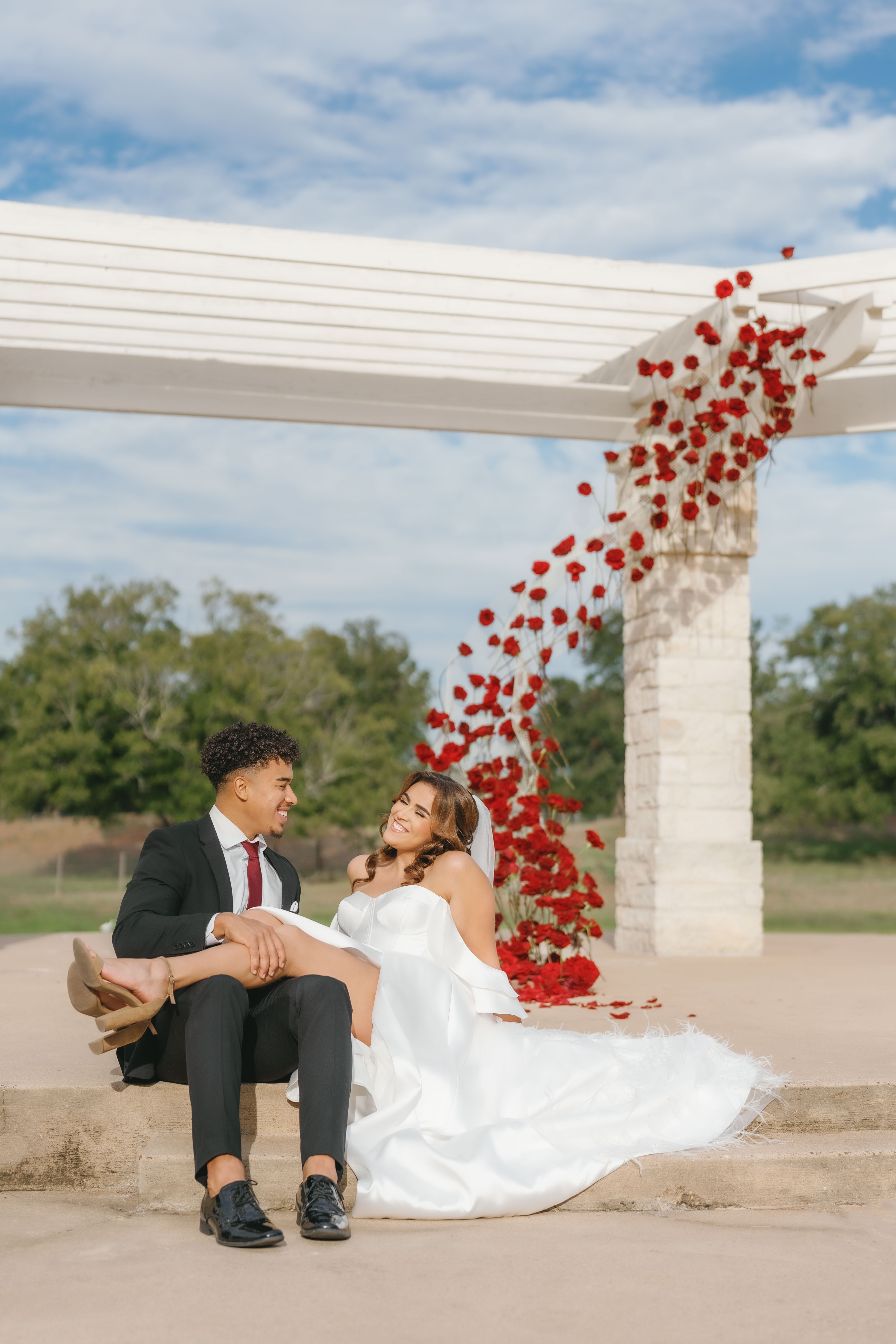 A smiling bride sits outdoors before a modern sculptural installation of vibrant red roses under a white arbor.