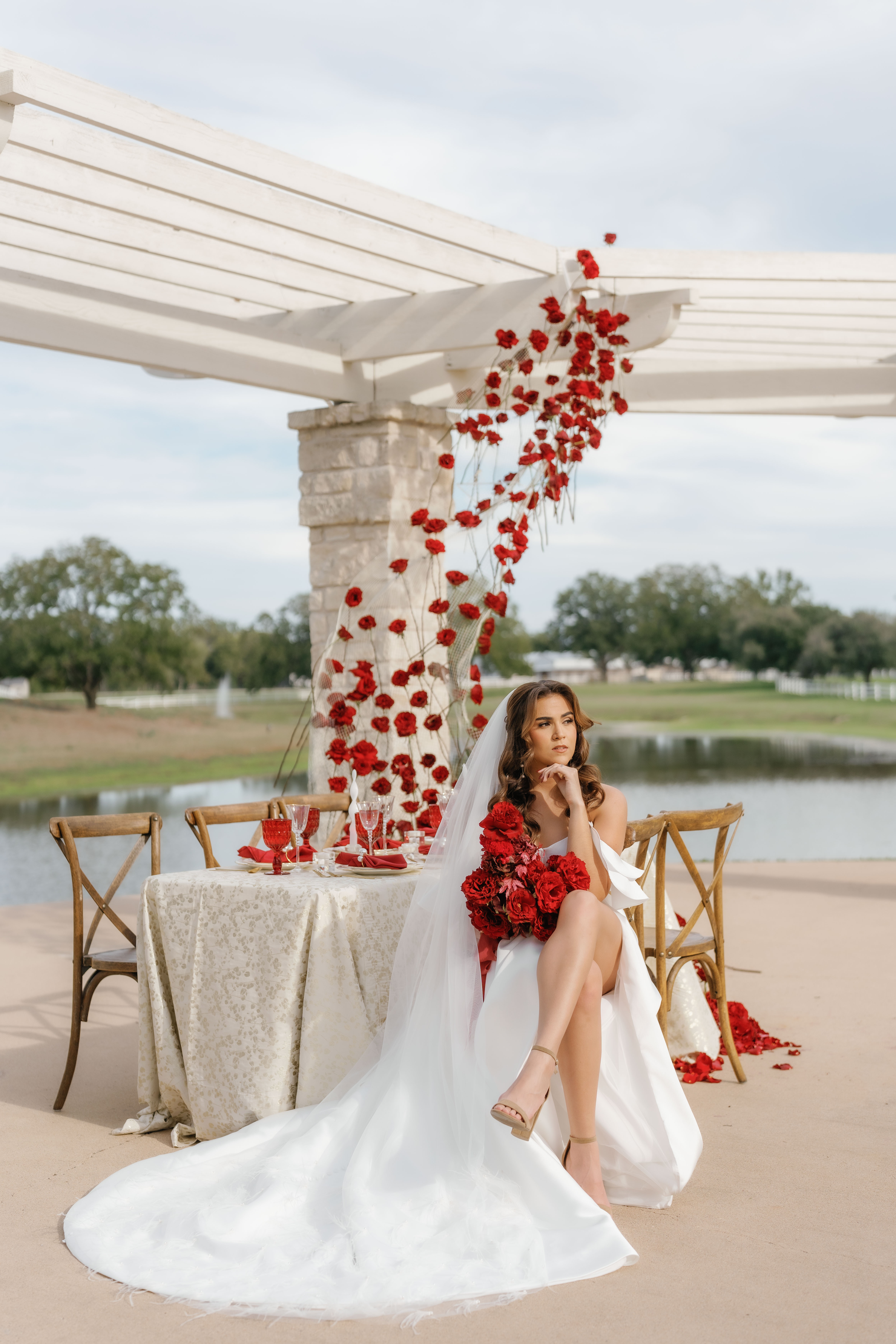 A bride in a white gown holds a red bouquet beside a sculptural red rose installation under an outdoor pergola.