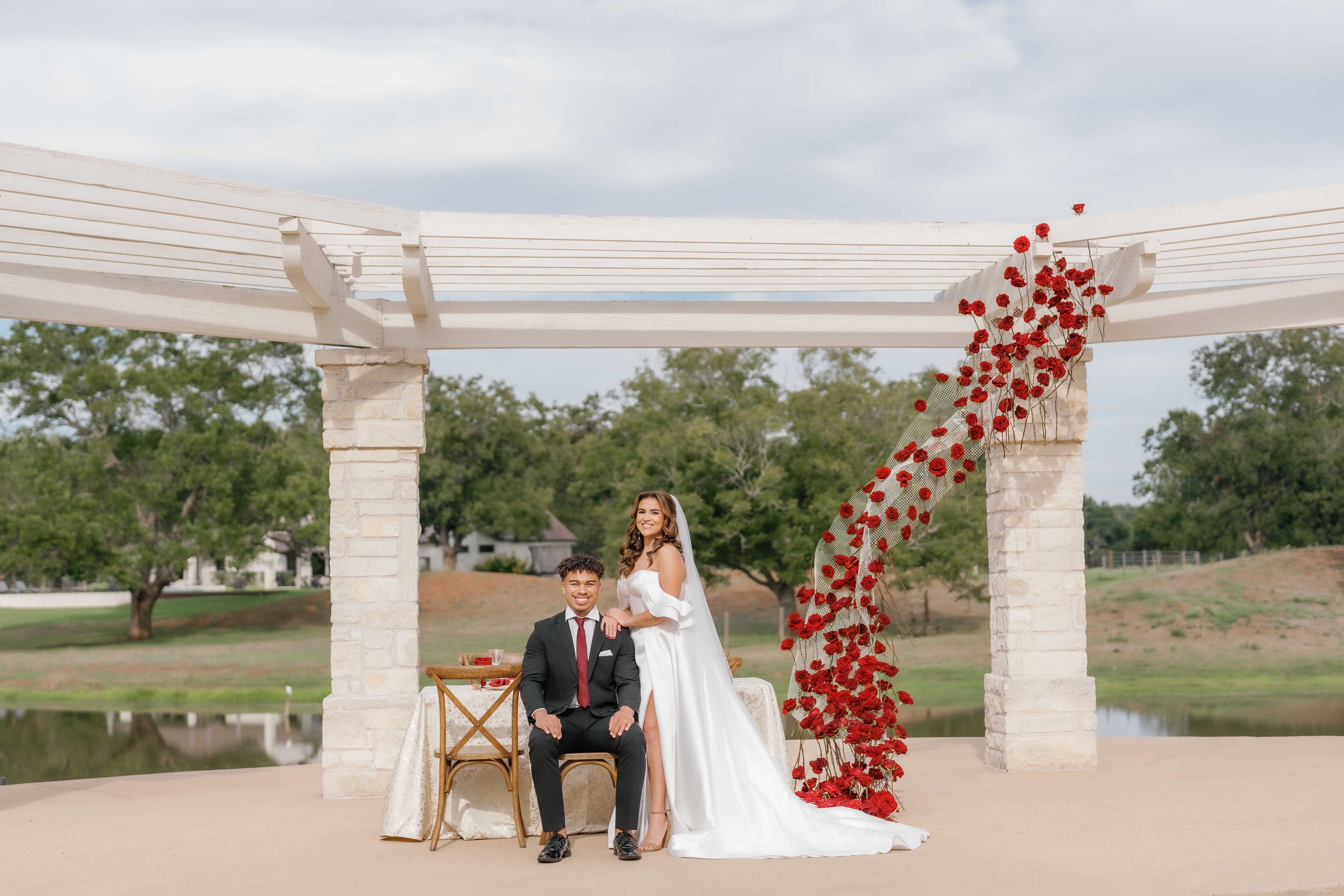A bride and groom pose together outdoors beneath a white pergola beside an installation of red roses.