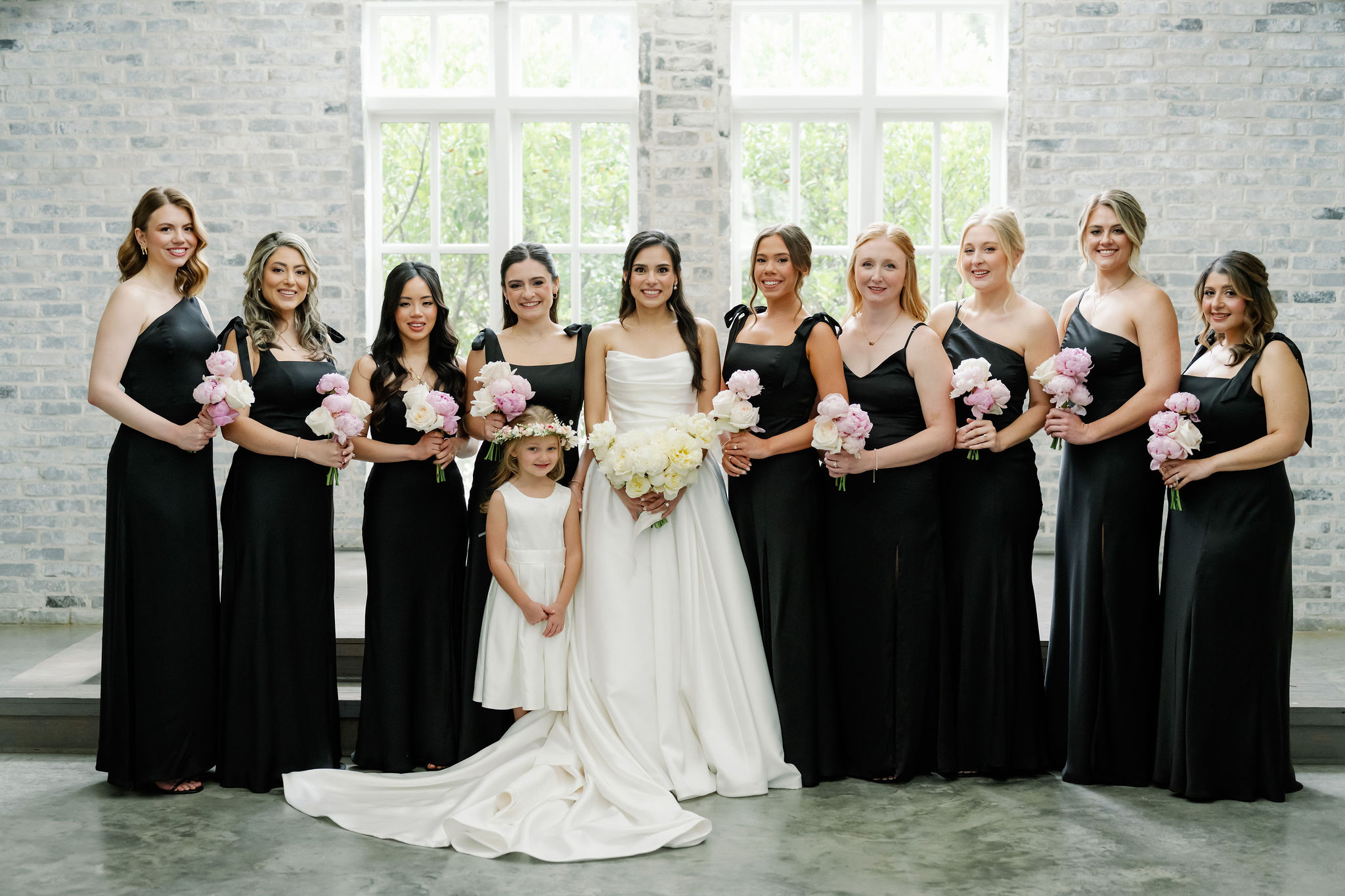 A bride in a strapless gown stands with bridesmaids in black holding soft pink bouquets and a flower girl with a floral crown.