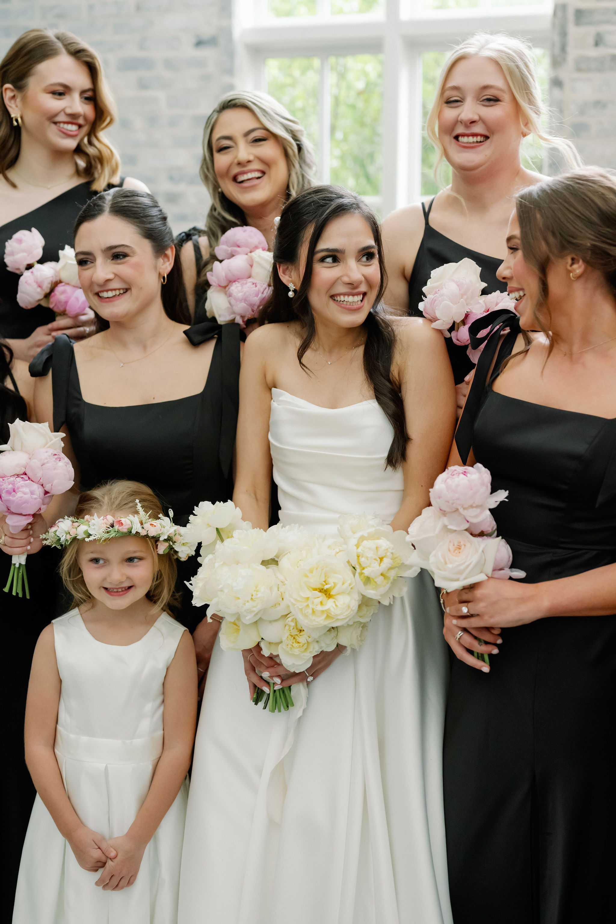 A bride holding an ivory peony bouquet poses with bridesmaids holding blush florals and a flower girl wearing a delicate floral crown.