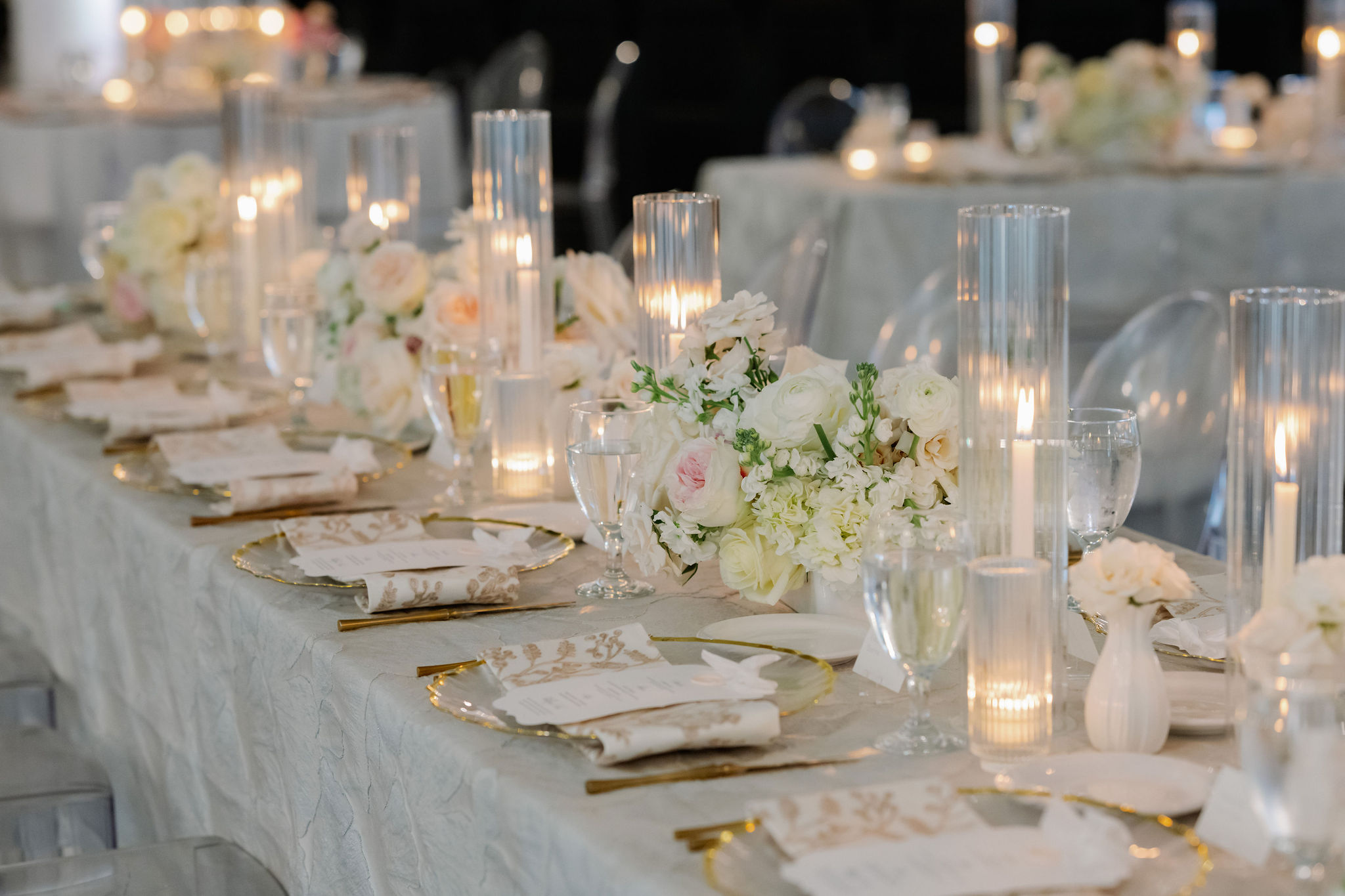 White and blush floral arrangements and glowing fluted glass candles line an elegant wedding reception table.