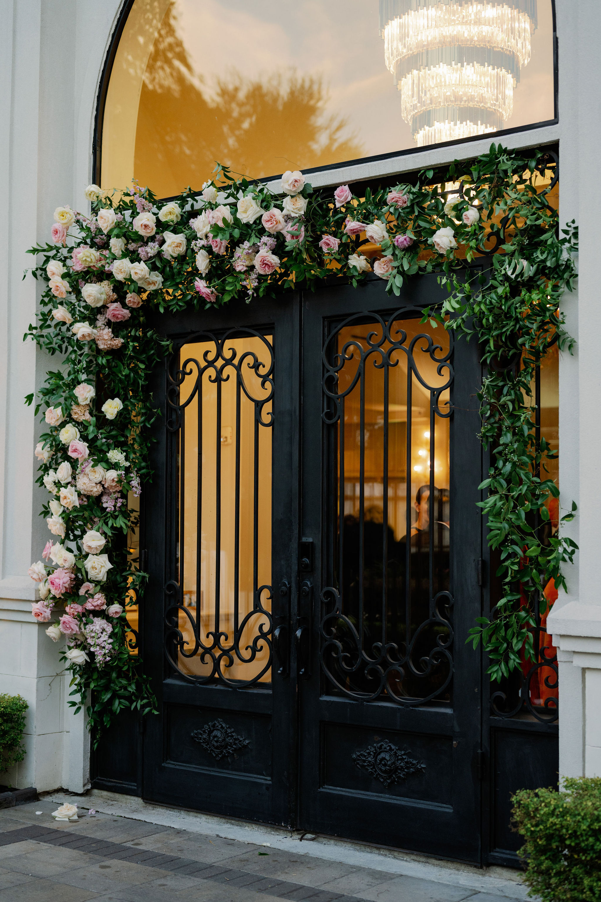 Lush pink and white floral garland frames grand black wrought-iron entrance doors beneath a crystal chandelier.