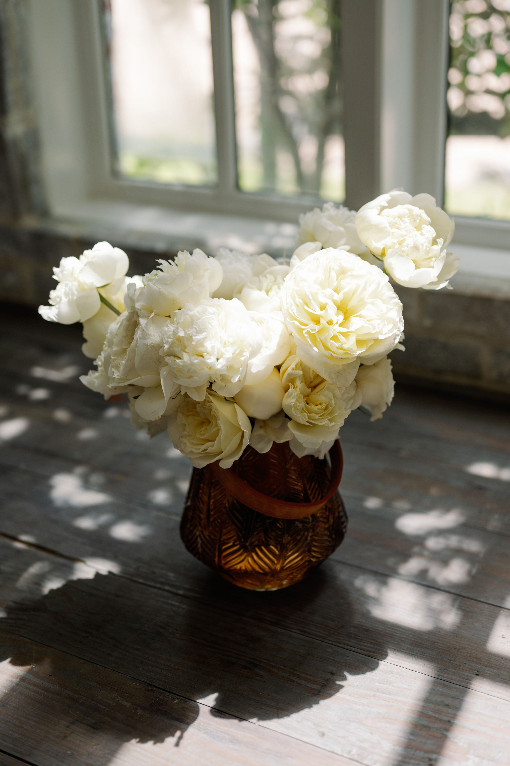 Lush white peonies and garden roses arranged in an amber fluted glass vase on a sunlit wooden table.