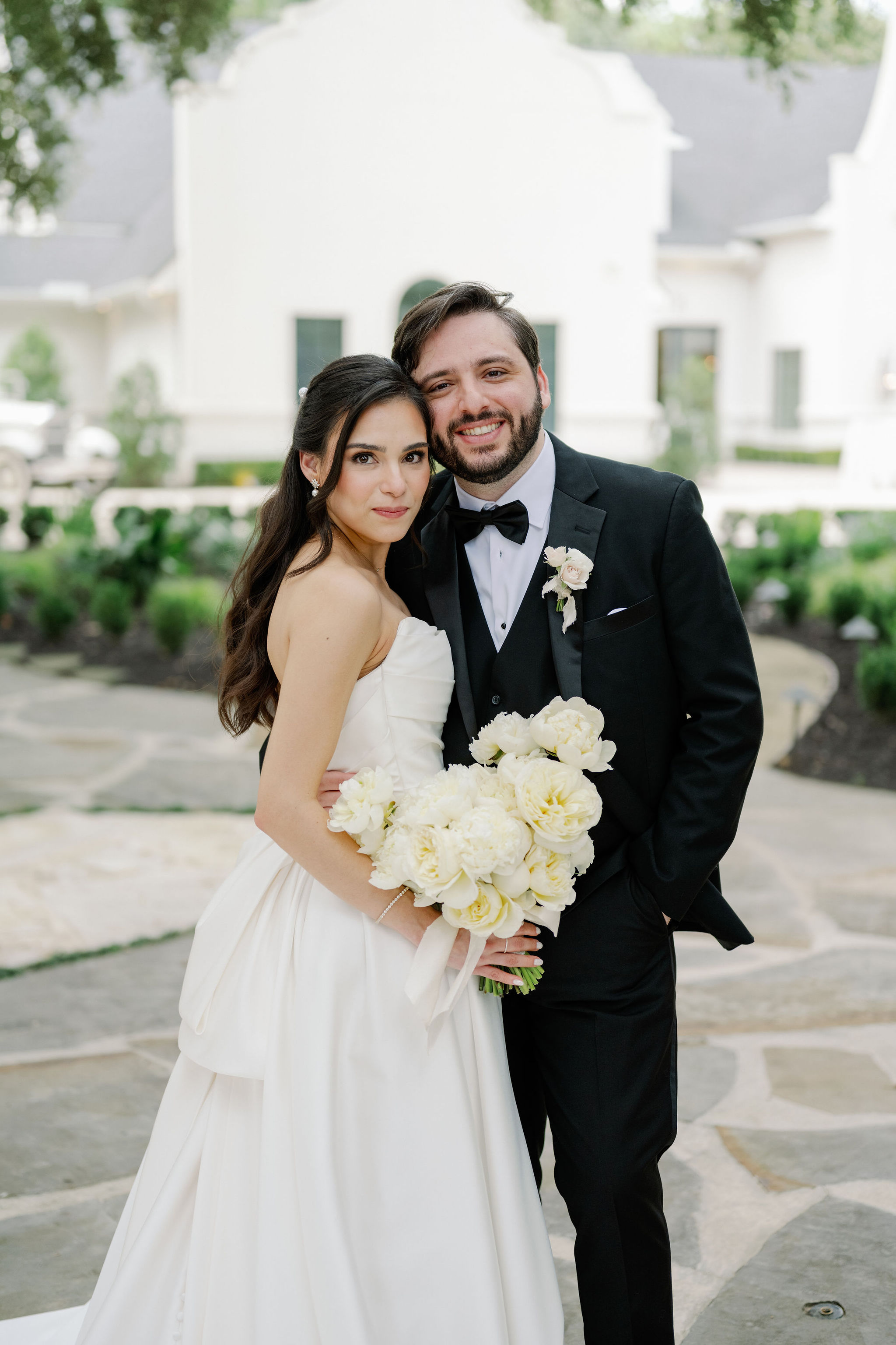 A bride in a strapless white gown holds a lush bouquet of ivory peonies alongside the groom in a classic black tuxedo.
