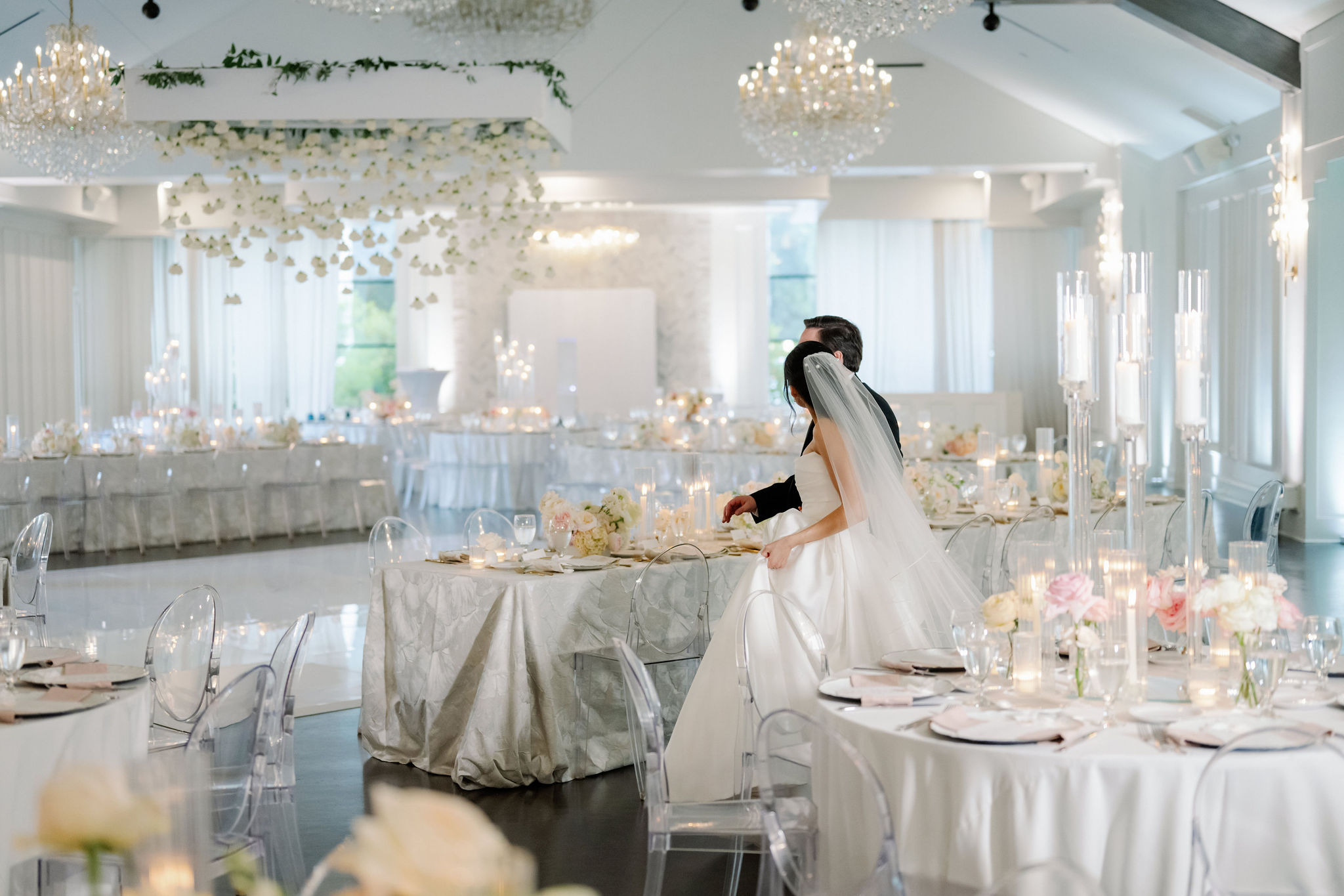 Bride and groom walking through a white ballroom reception with candlelit tables and crystal chandeliers.