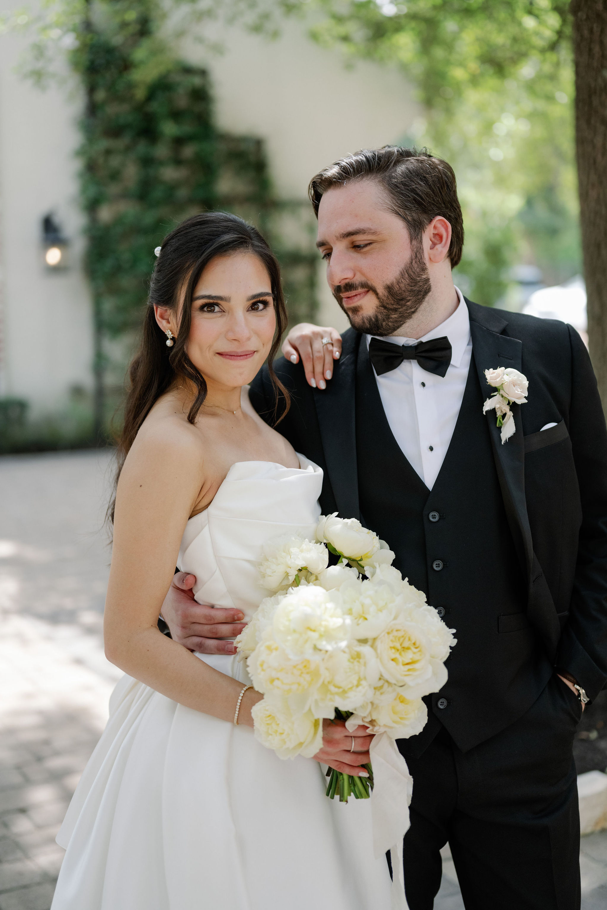 A bride in a strapless gown holds a white peony bouquet while embracing a groom in a black tuxedo.