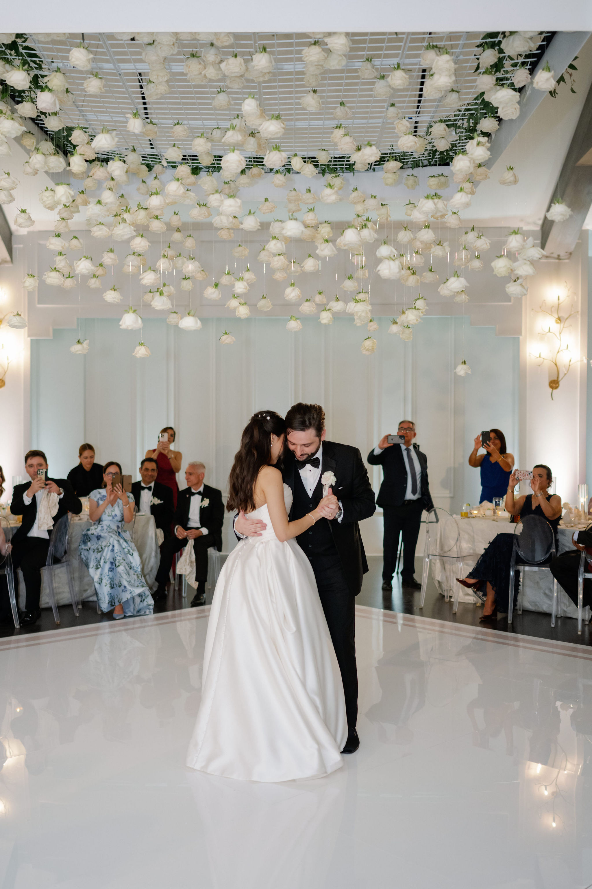 A bride and groom dance on a white dance floor beneath a suspended installation of hanging white roses.