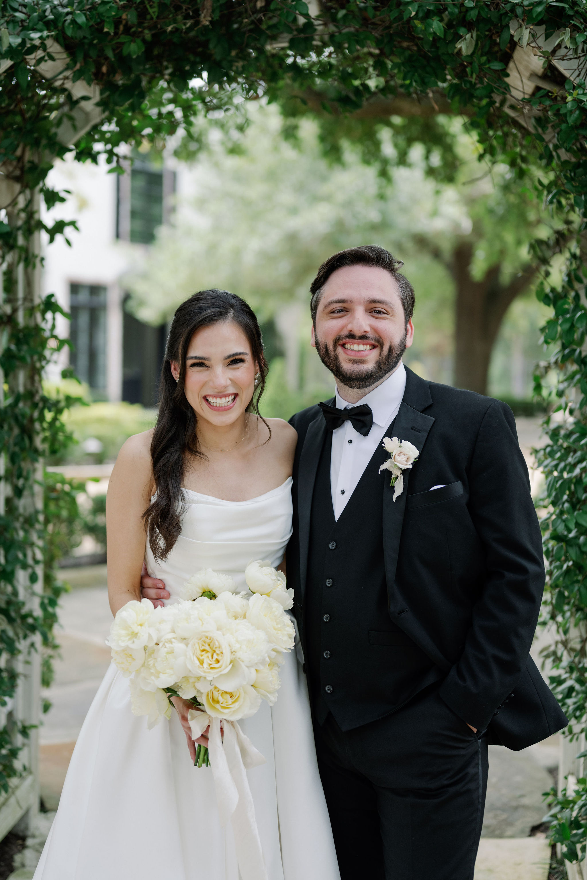 Smiling bride in a strapless gown holding a lush white floral bouquet beside the groom in a classic black tuxedo.