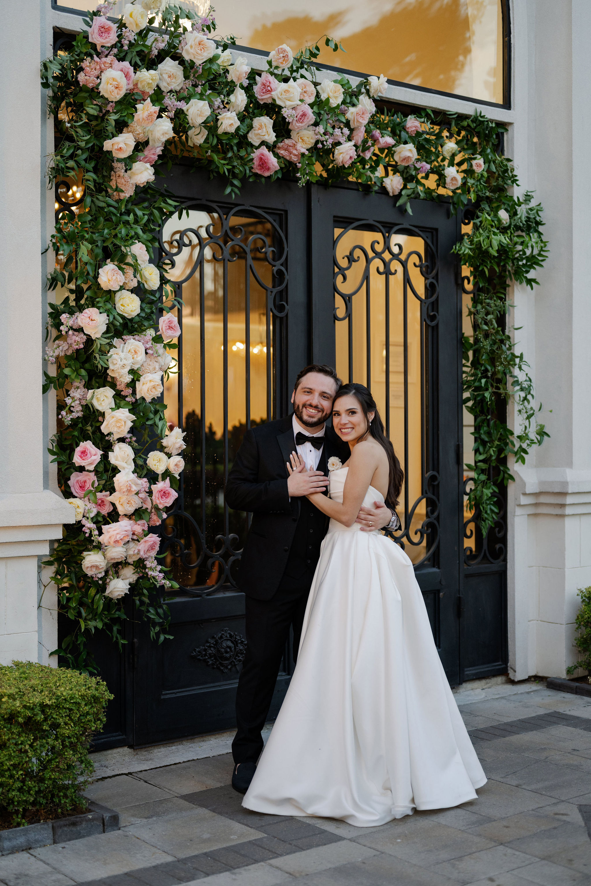 Bride and groom smiling together in front of ornate wrought-iron doors decorated with lush pink and cream roses.