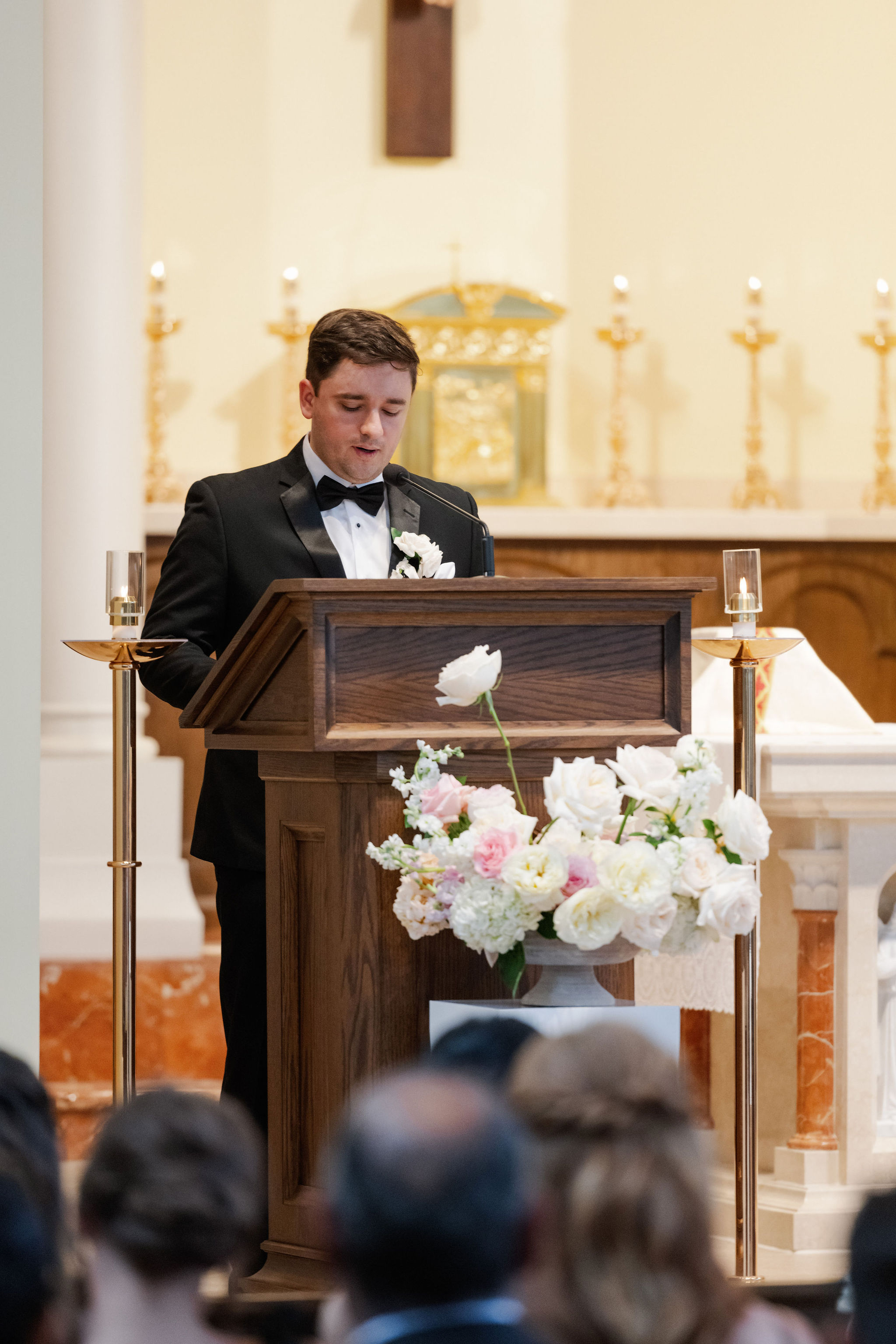 A man in a tuxedo speaks at a wooden lectern beside a white and blush floral arrangement during a wedding ceremony.